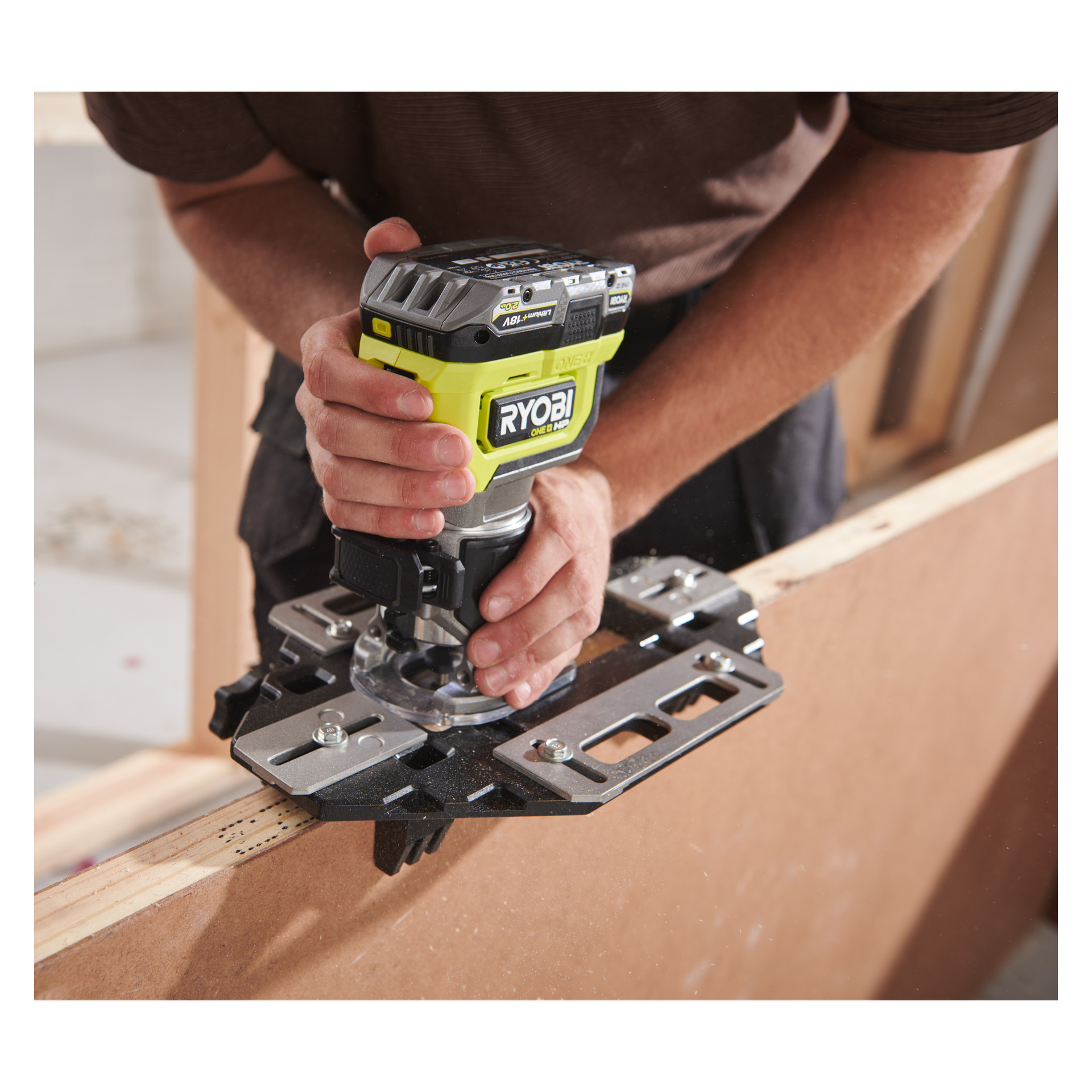 Man griper en Ryobi One+ HP håndholdt ruter RTR18C-0 med begge hender for å frosse kanten av treverket på en arbeidsplass.