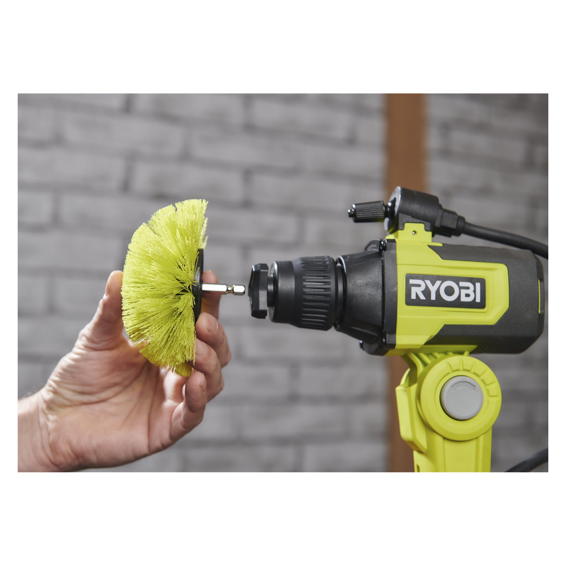 Någon kopplar en Ryobi USB Lithium Cleaning-borste RACLRM110 till en borrmaskin, med en grå tegelvägg bakom sig.