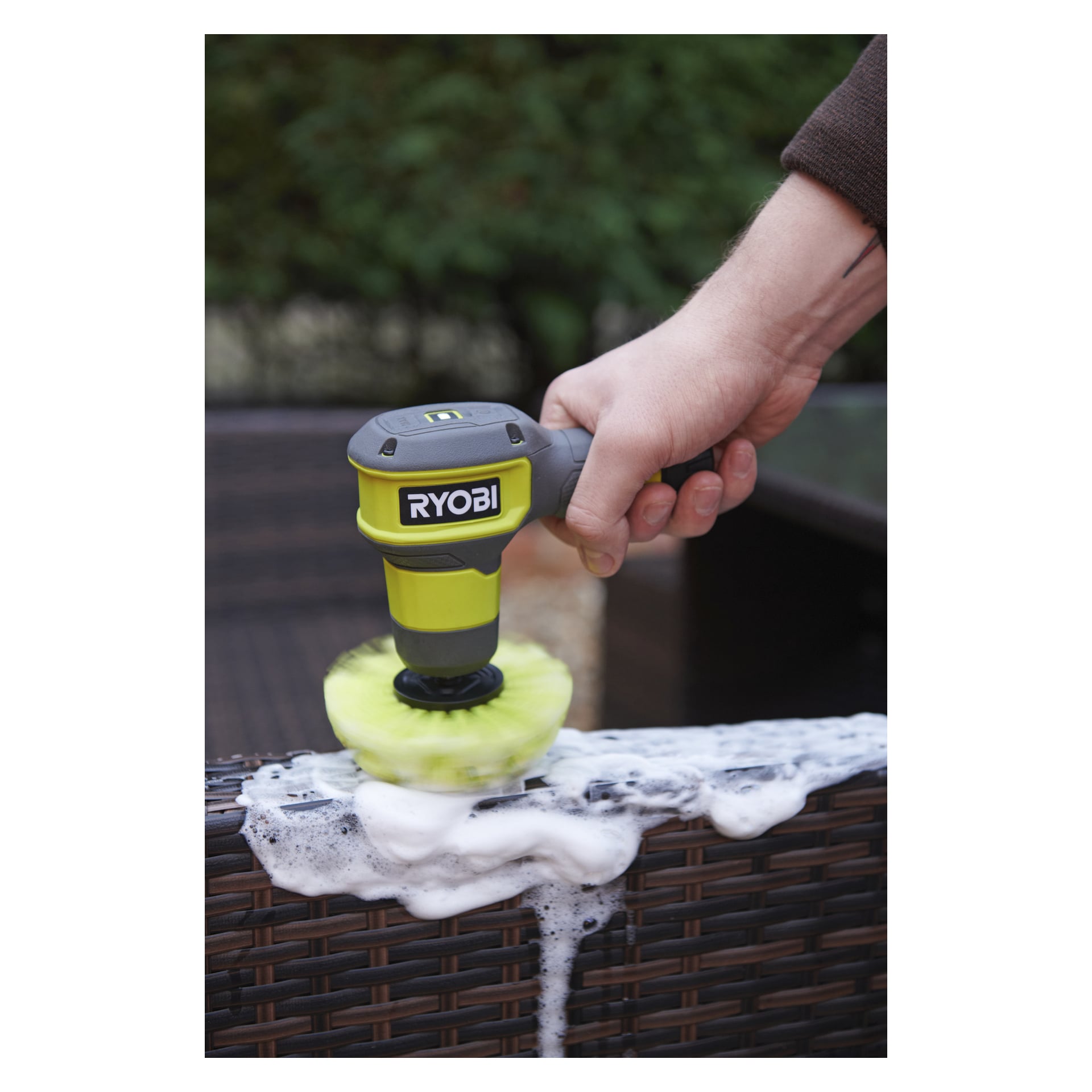 En Ryobi USB Lithium Cleaning borste med gult huvud skrubbar tvålbad, grönt löv mjukt suddigt i bakgrunden.
