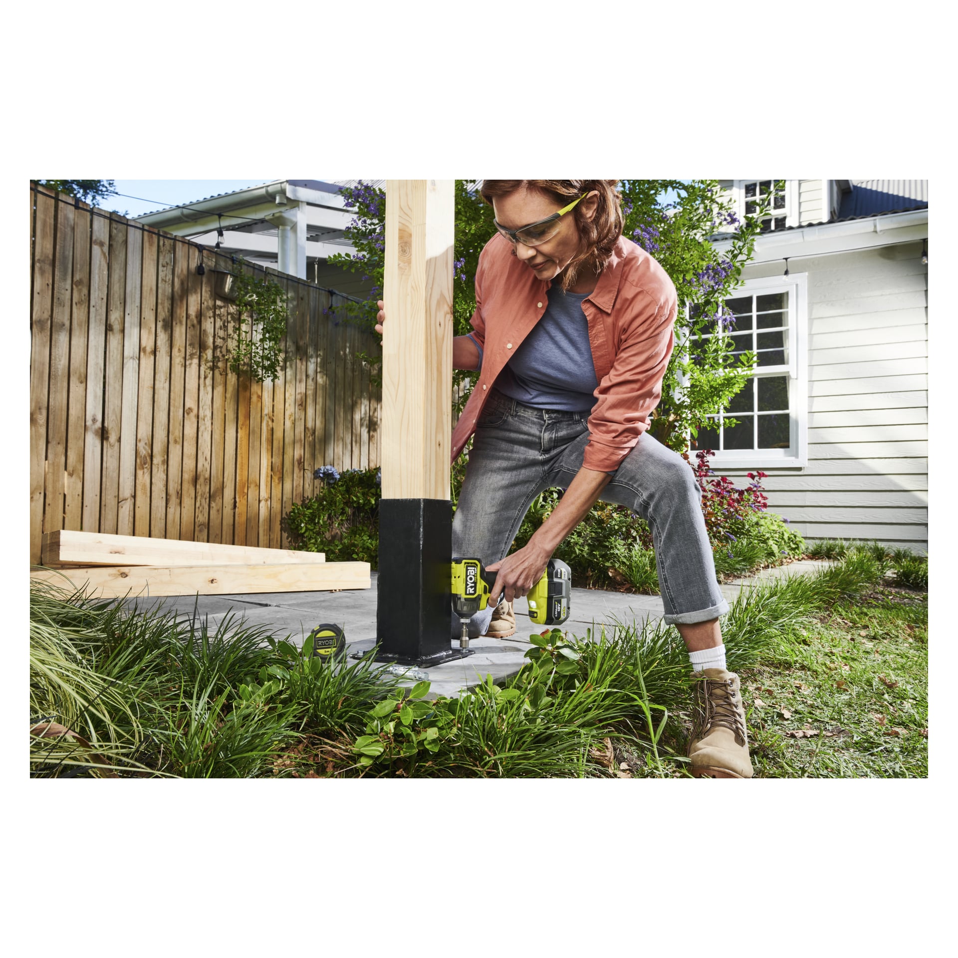 En person med skyddsglasögon och stövlar använder en Ryobi One+ HP Impact Driver för att fästa en trästolpe i betongen utomhus, med verktyg i närheten.