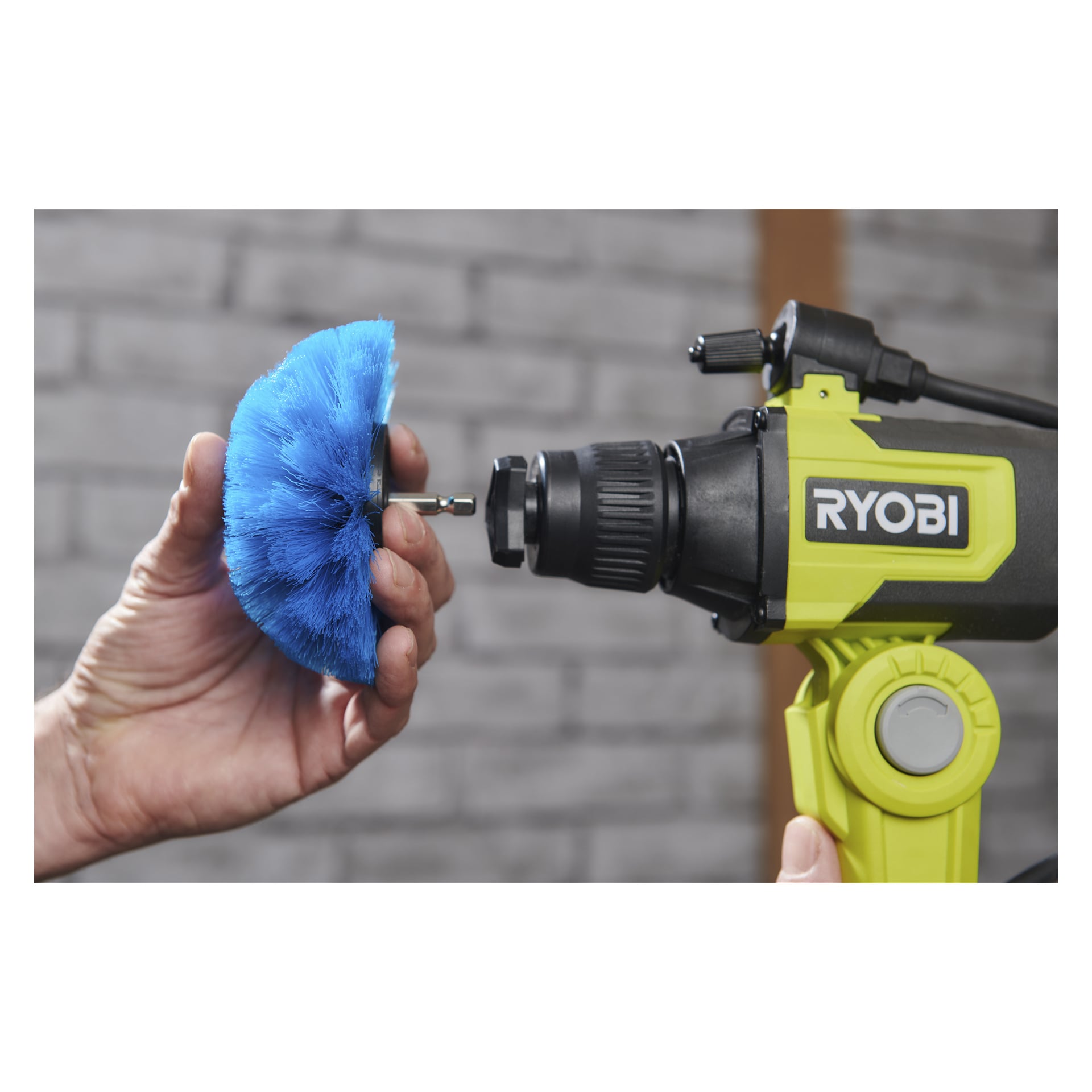 Någon fäster en blå Ryobi USB Lithium RACLRS110 rengöringsborste på en gul och svart borr; Den grå tegelväggen är suddig bakom.