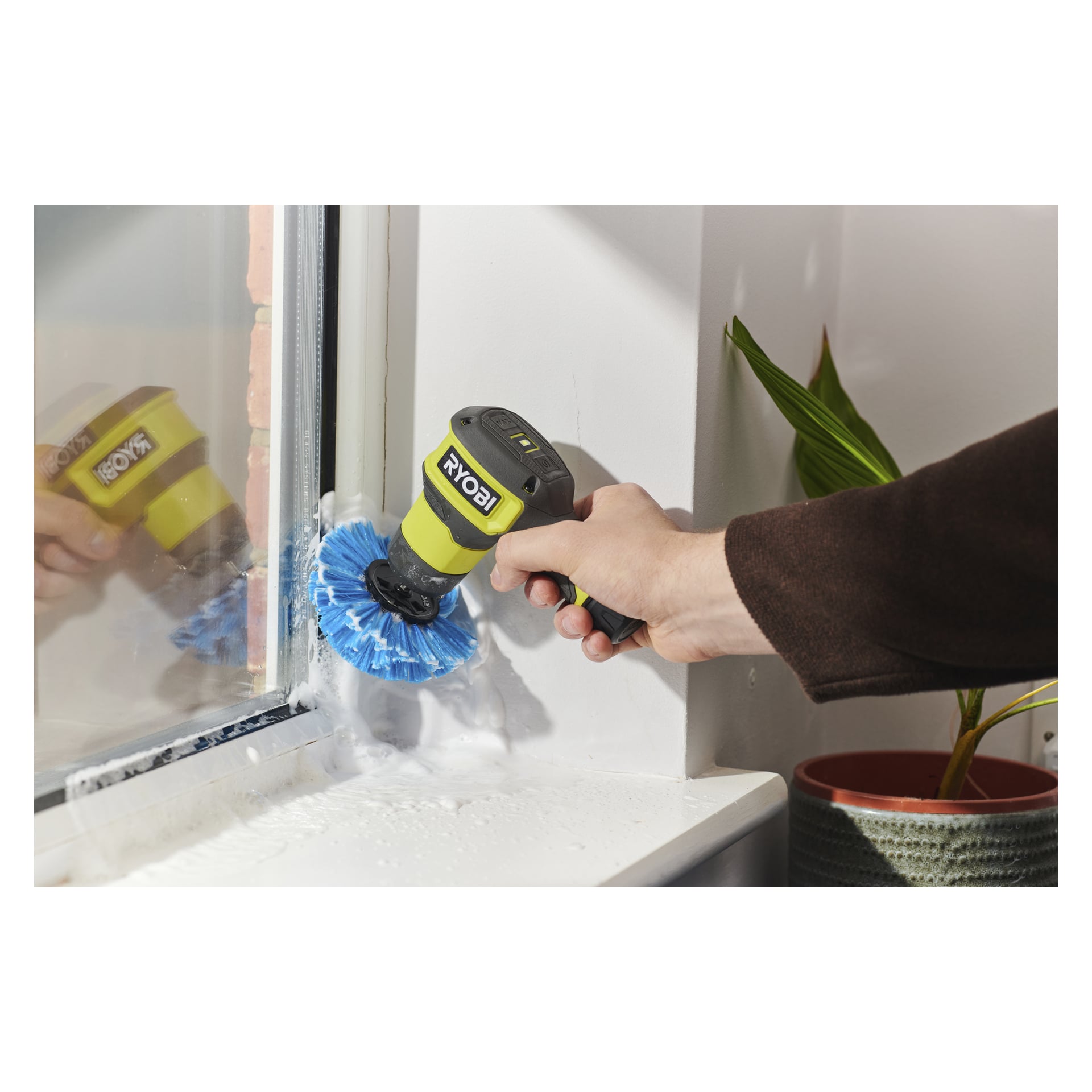 En person rengör ett hörn i fönsterkarmen med en Ryobi USB Lithium Cleaning-borste och ett blått huvud; tvålskum och en grön växt i närheten.