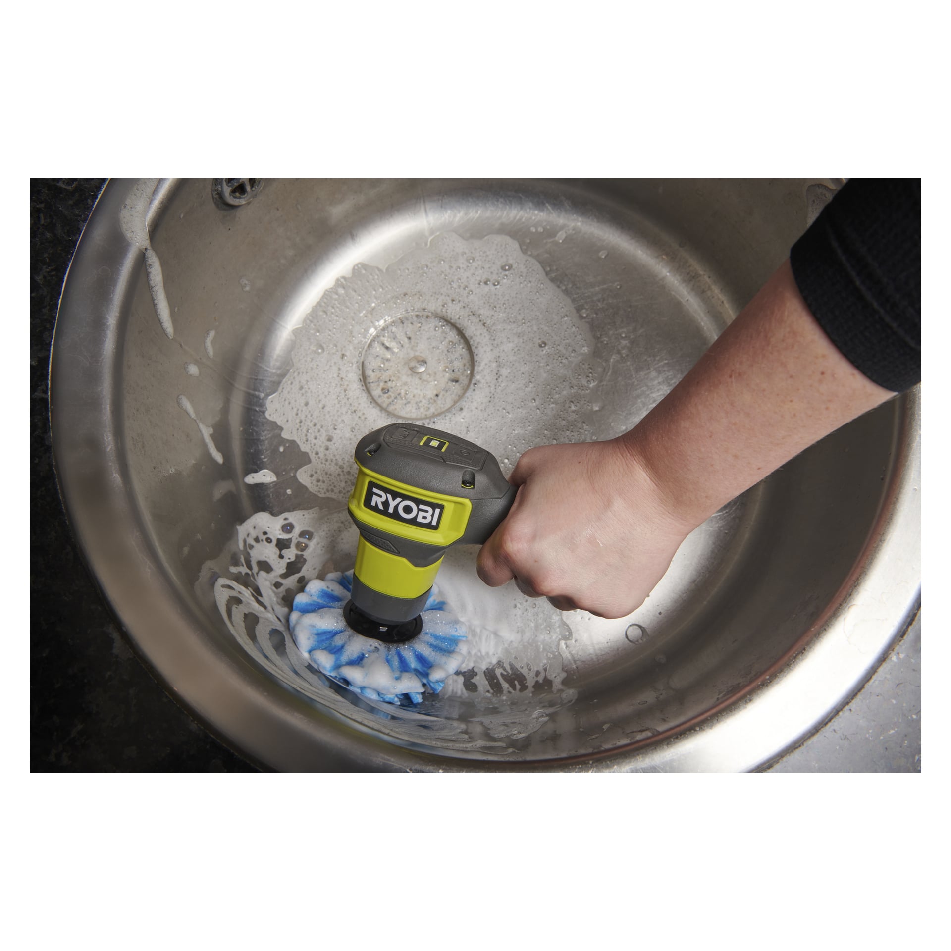 Någon använder en Ryobi USB Lithium Cleaning Brush RACLRS110 med ett blått och vitt tillbehör för att tvätta en diskho i rostfritt stål.