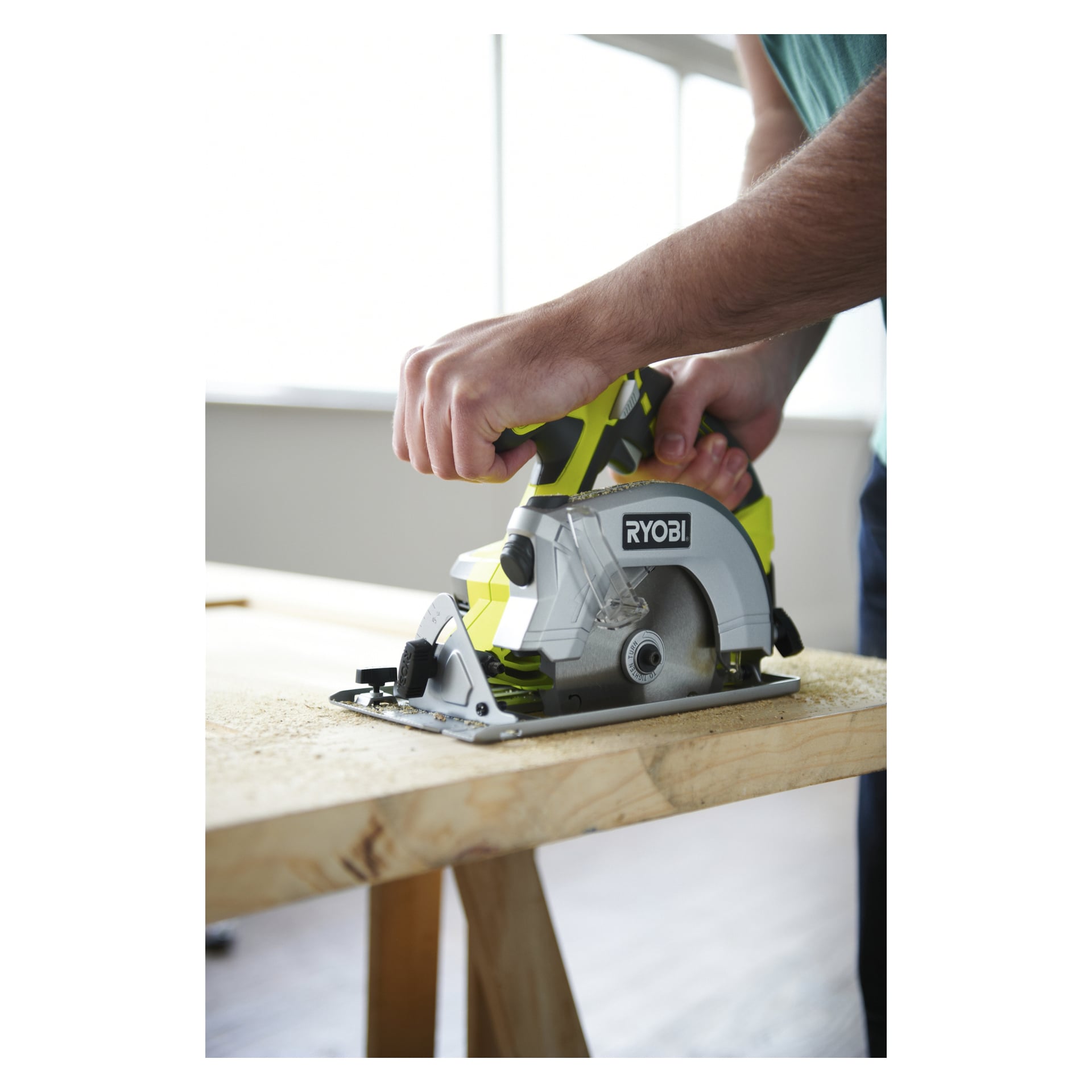 Någon använder en Ryobi One+ cirkelsåg R1801WSLN för att såga trä på en arbetsbänk i en ljus inomhusverkstad.