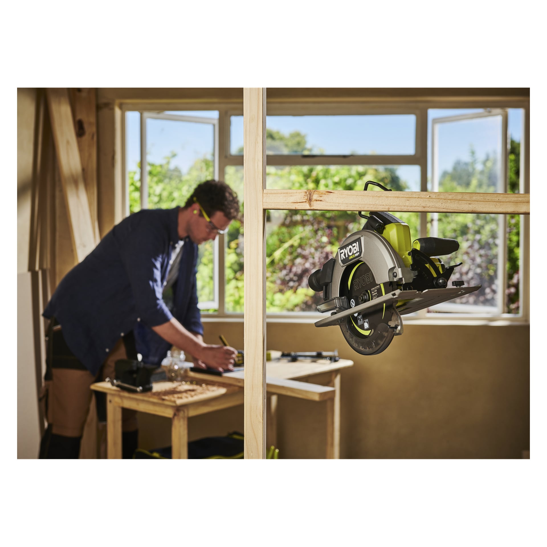 En person jobber ved et bord nær store åpne vinduer, med en Ryobi One+ HP sirkelsag RCS18X1-0 hengende fra en treramme foran.