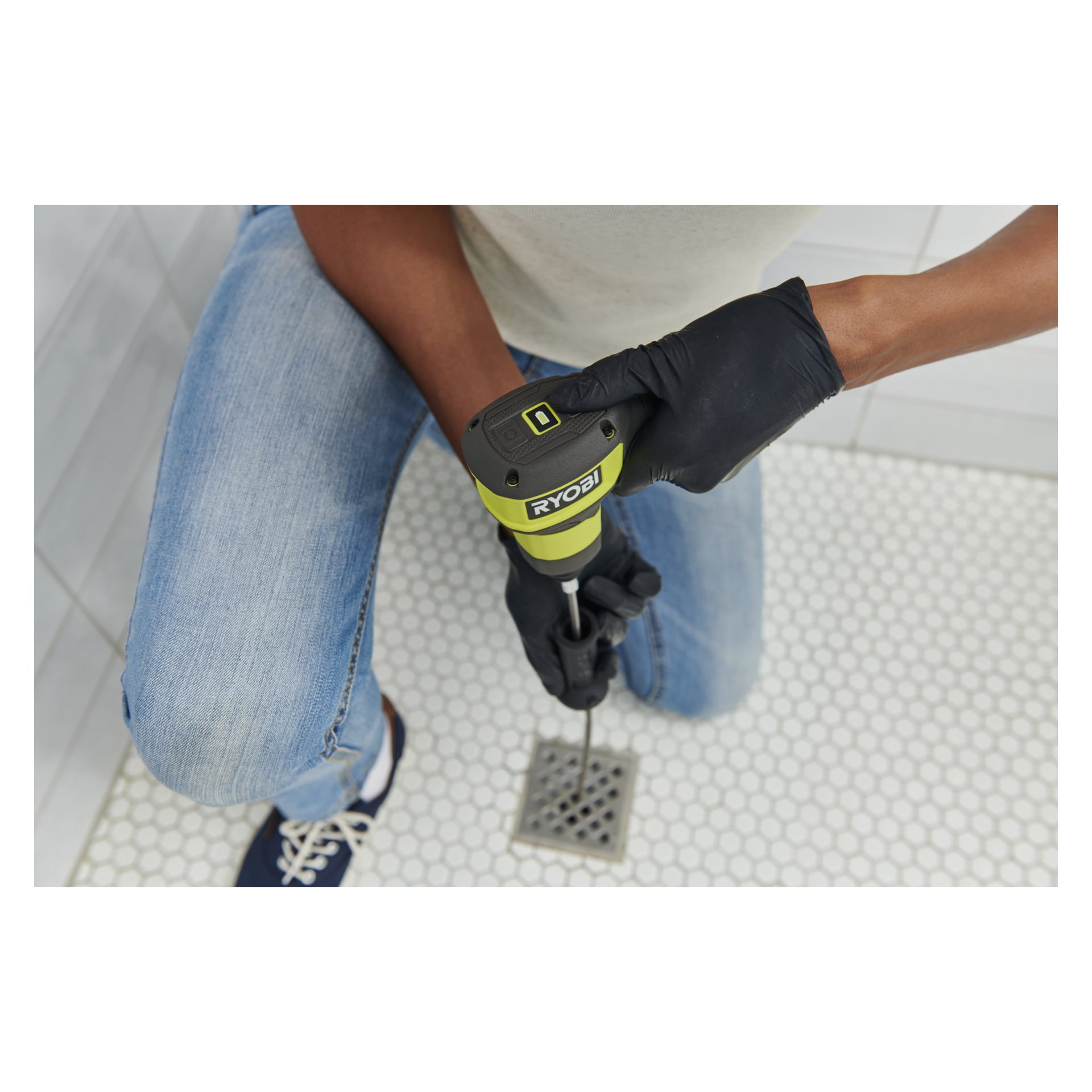En person i svarte hansker og jeans bruker en Ryobi USB Lithium avløpsrens for å åpne et metallavløp på hvite sekskantede fliser.