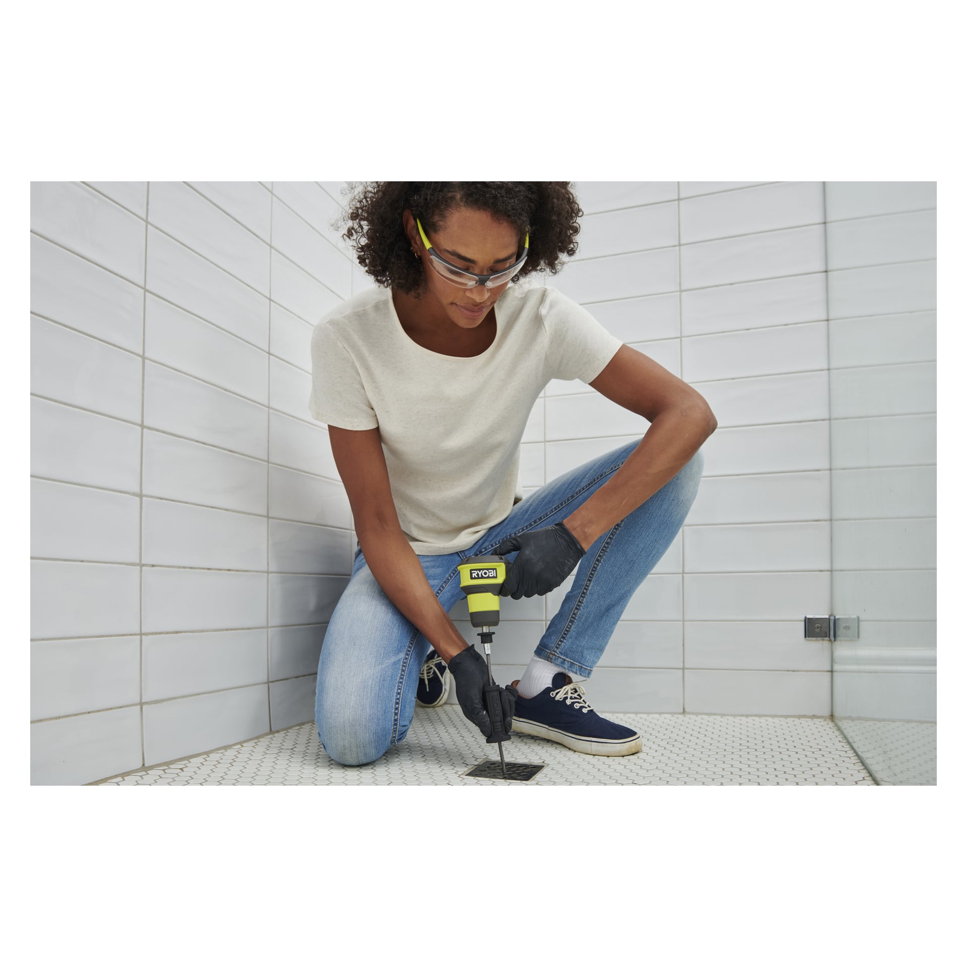 En person med vernebriller, hansker, hvit t-skjorte og jeans bruker en Ryobi USB Lithium avløpsrenser på et flislagt gulvavløp.