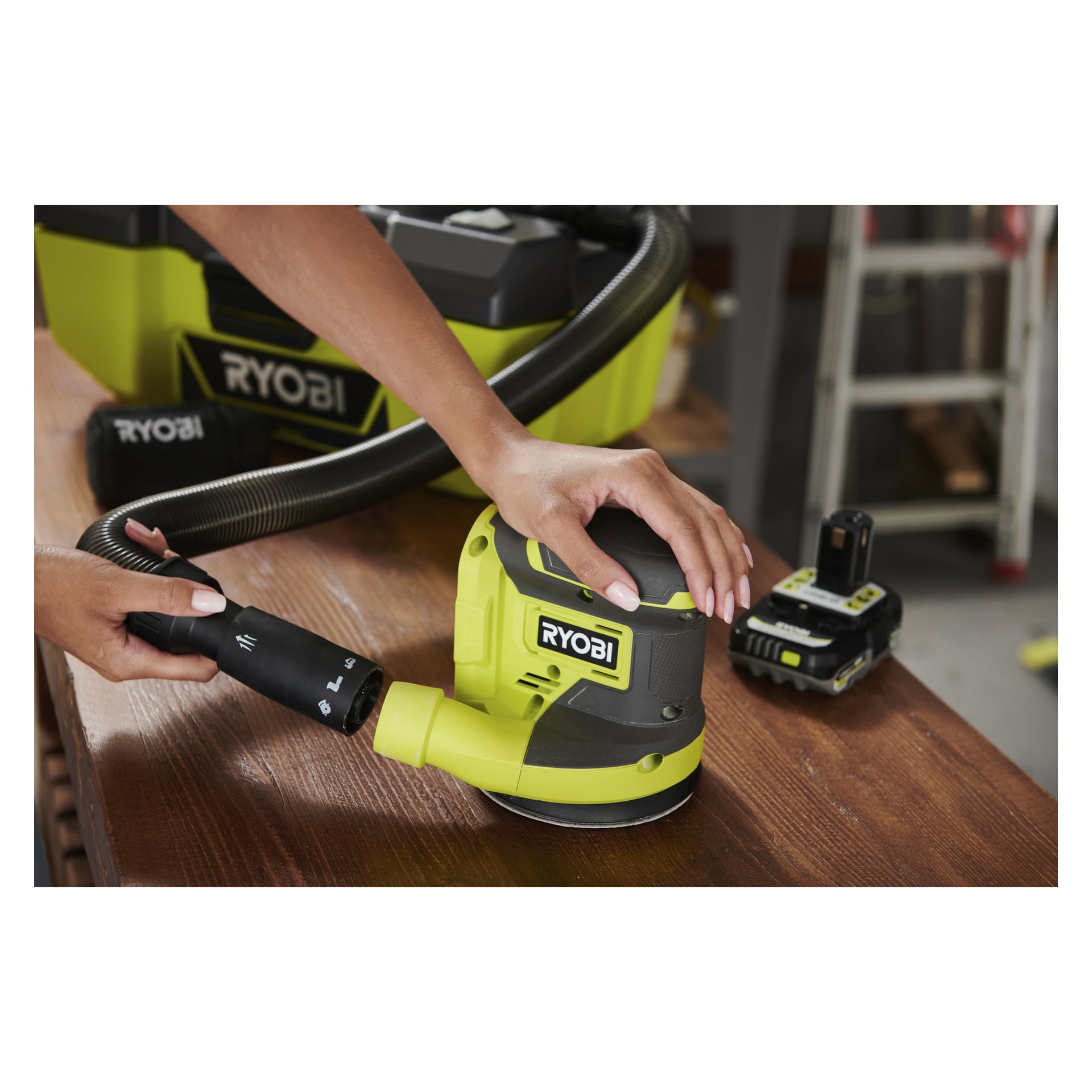 Ktoś szlifuje drewno szlifierką Ryobi One+ Eccentric R18ROSN-0, podłączoną do niej podciśnieniową wąż, z baterią i walizką narzędziową z tyłu.