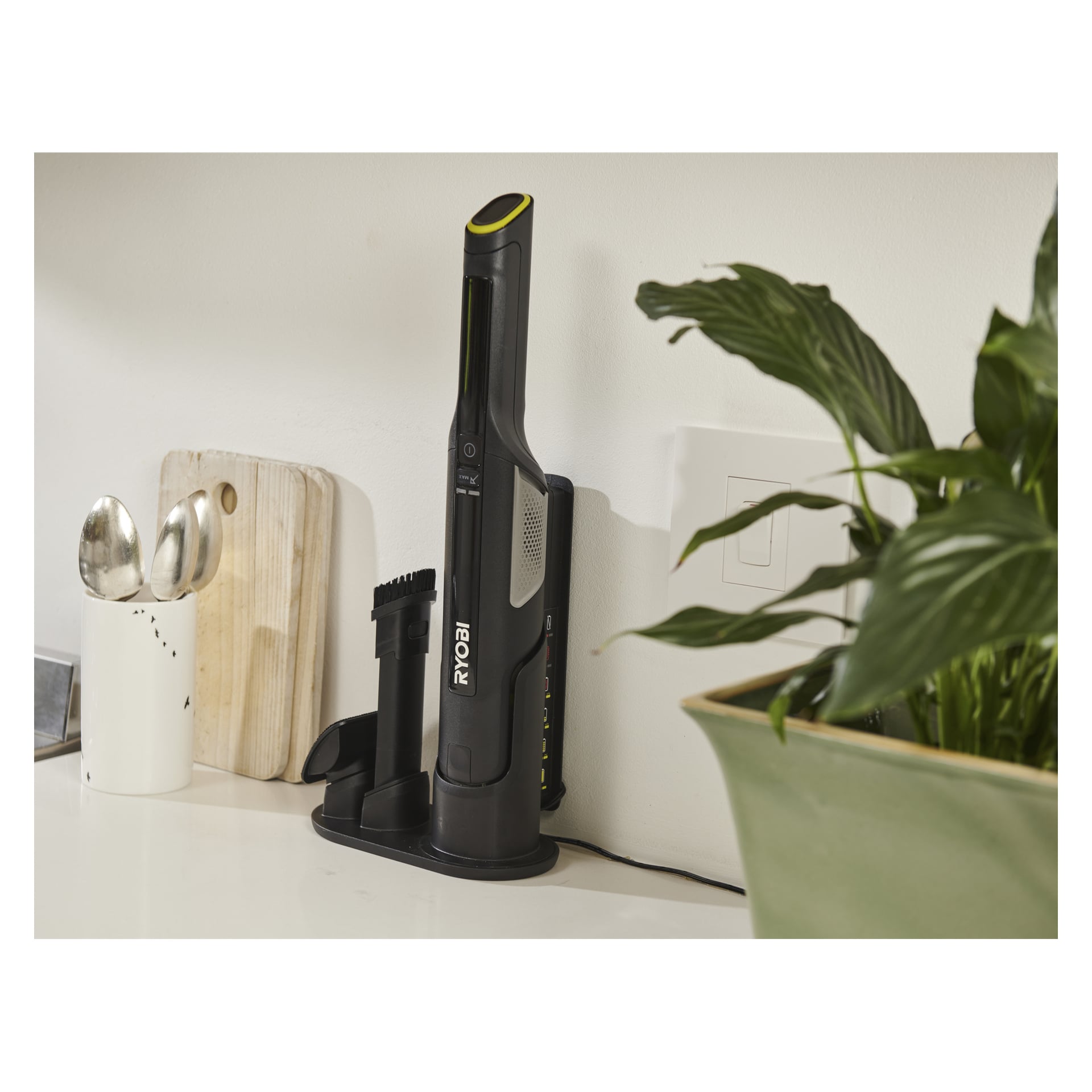 En Ryobi USB Lithium Handhållen dammsugare RHV44-220G laddas i sin docka på en köksbänk nära en skärbräda, skedar och grön växt.