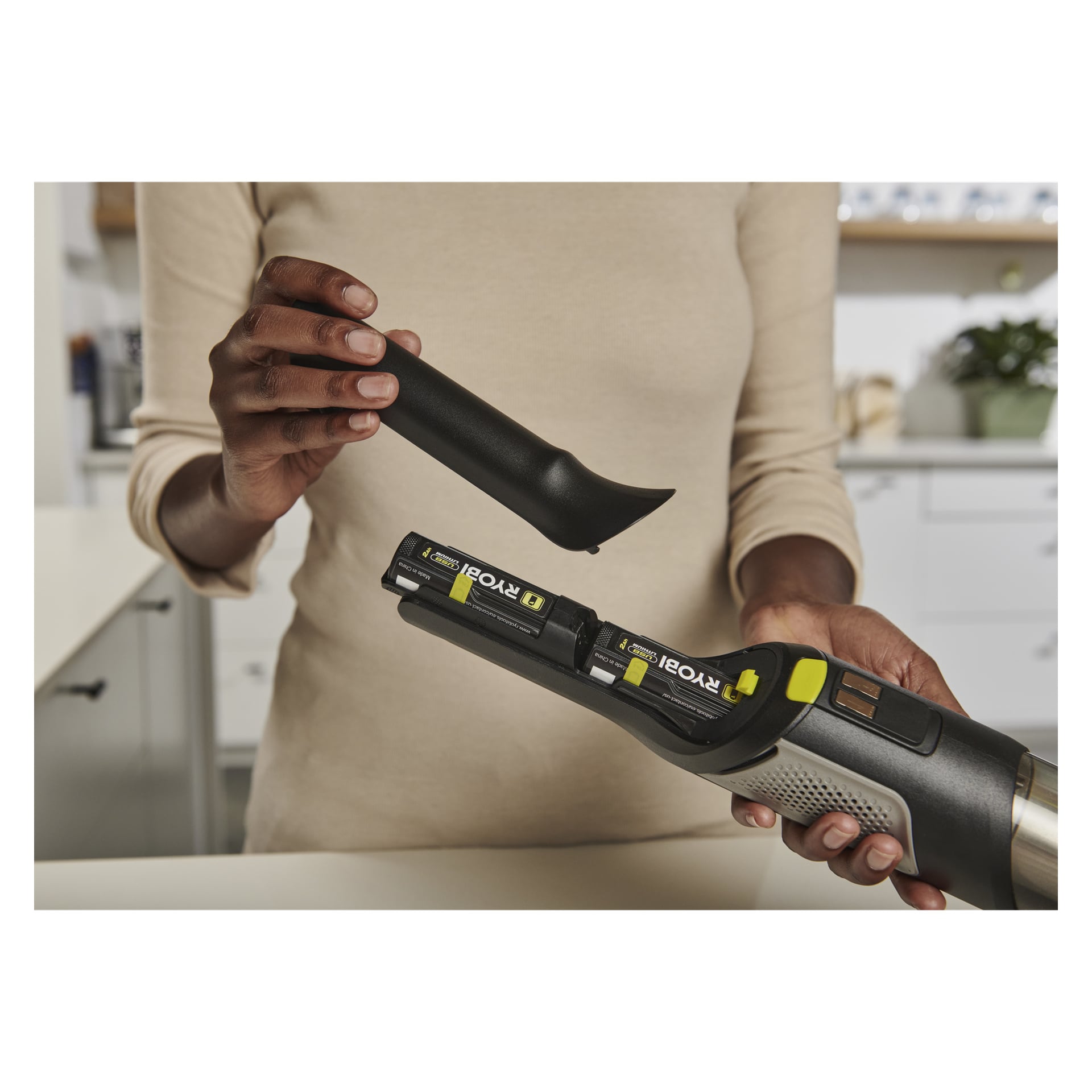 En person i ett modernt kök placerar två Ryobi USB Lithium AA-batterier i RHV44-220G handhållna dammsugare medan man håller i munstuket.