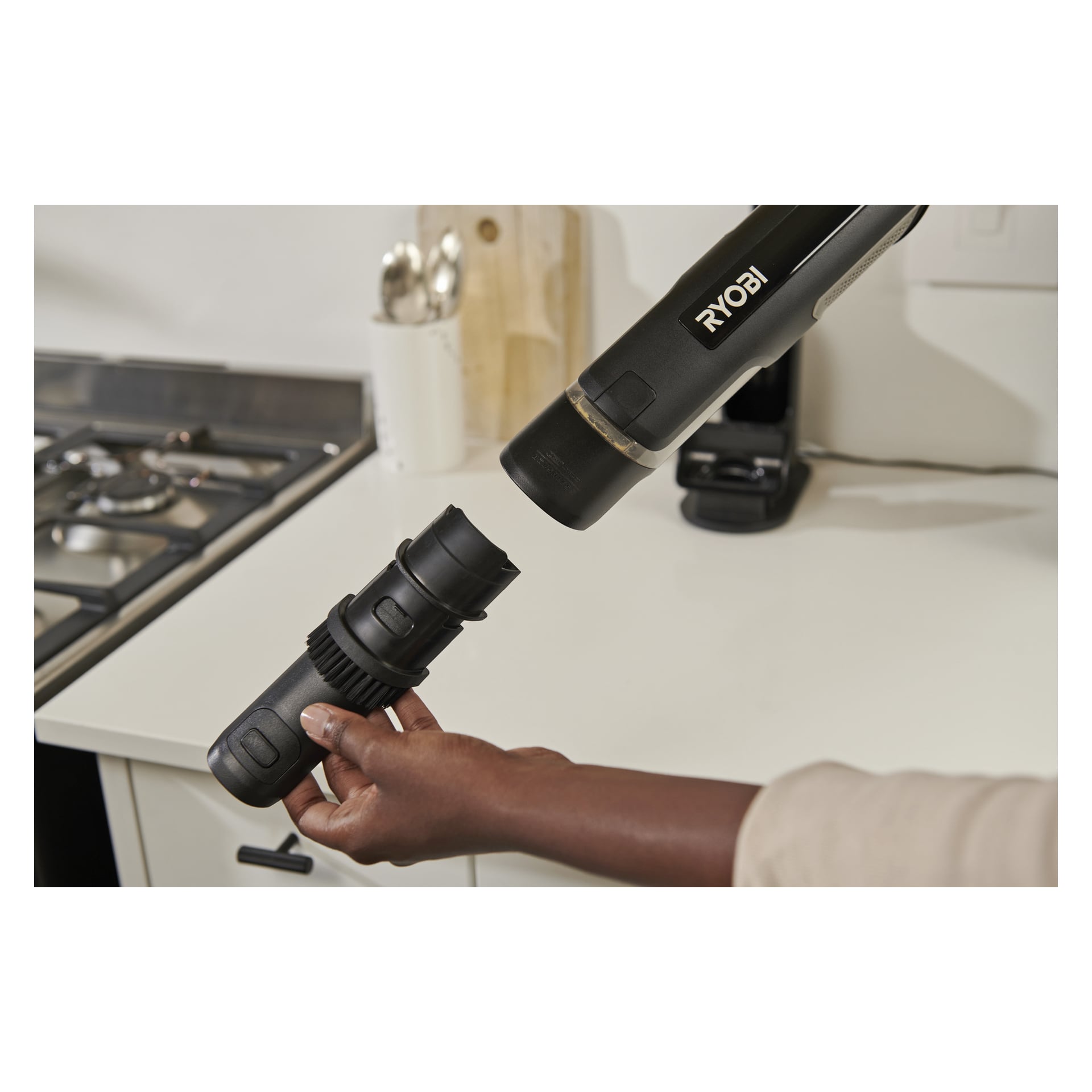 En person kopplar ett munstycke till Ryobi USB Lithium Handheld Dammsugare i ett kök med spis, redskap och laddare i närheten.