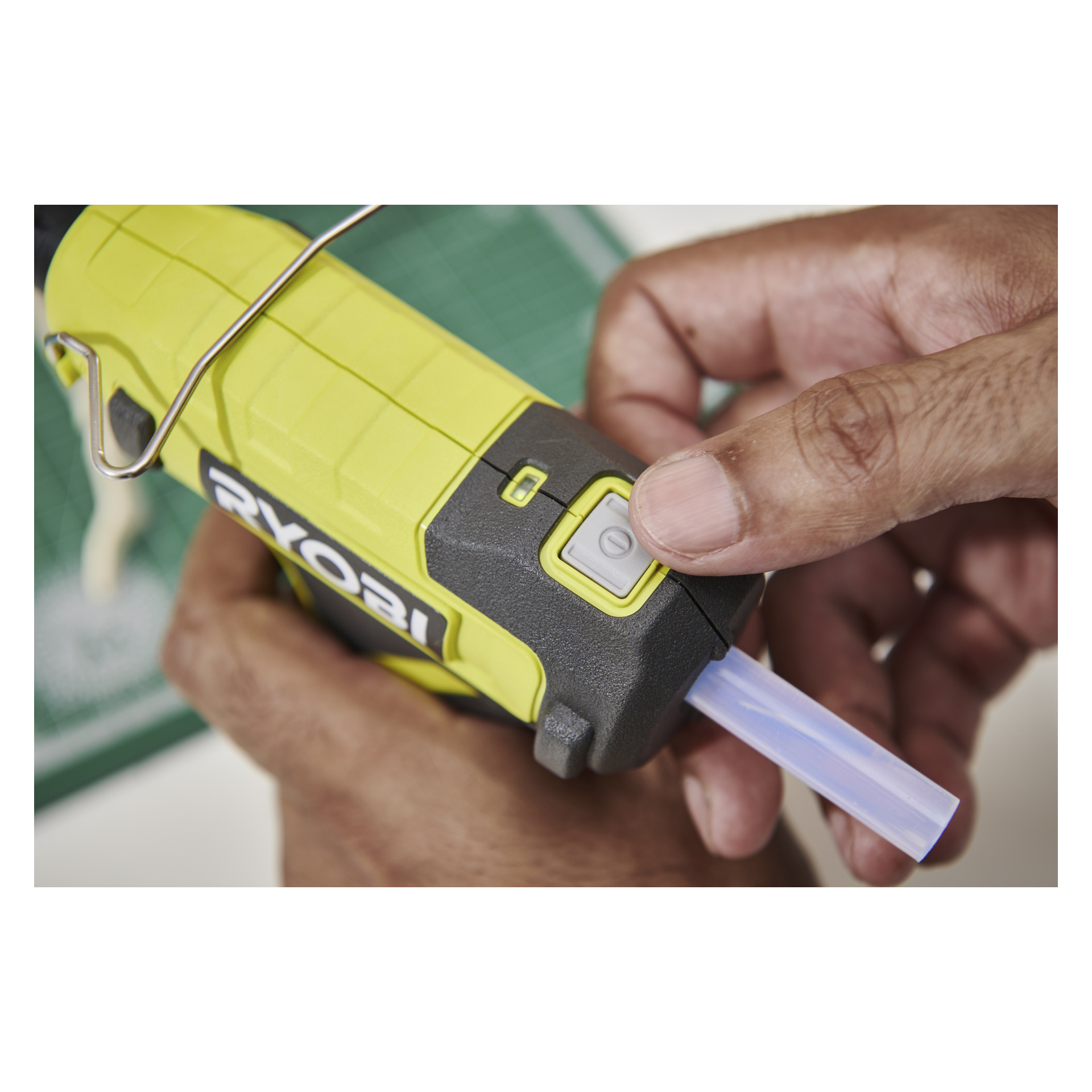 Ręka włącza pistolet Ryobi USB Lithium Battery RGLU4-0 z klejem w sztyfcie, ustawiony na zielonej matie do cięcia.