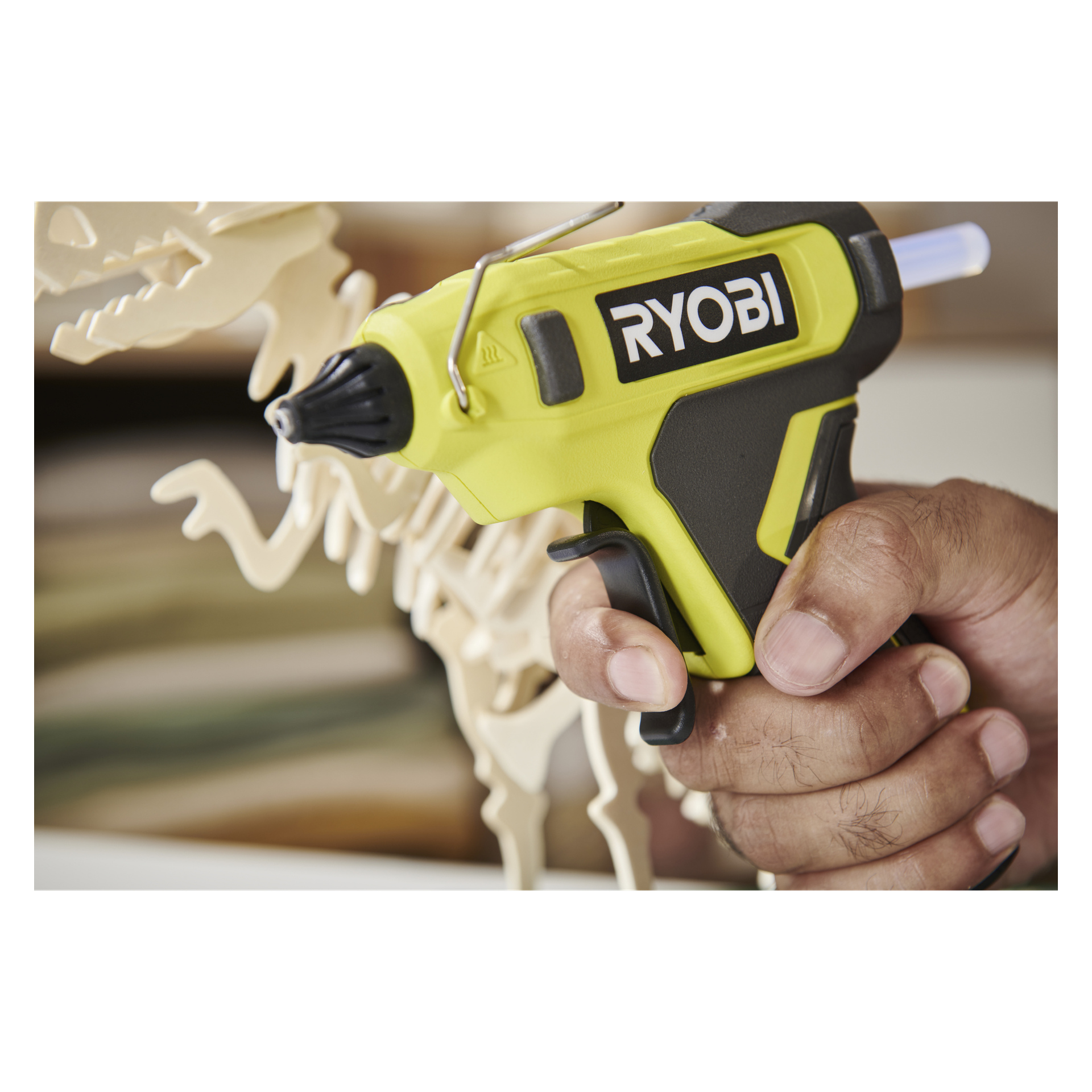 Ktoś trzyma żółto-pistolet Ryobi USB Lithium Glue i kieruje go w stronę drewnianego modelu dinozaura.