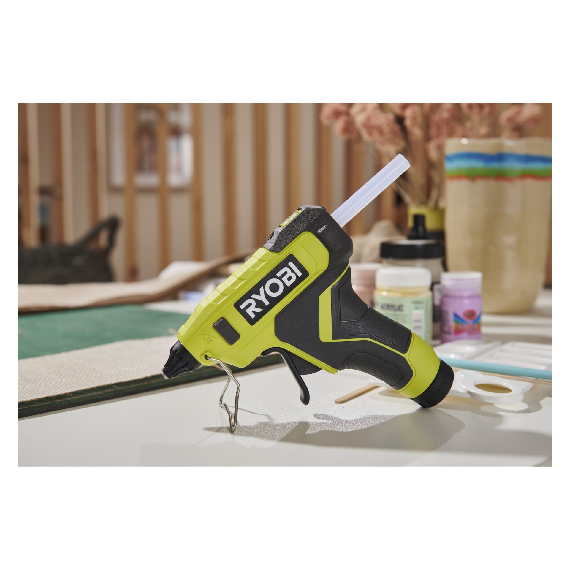 Na stole stoi pistolet Ryobi RGLU4-0 USB z klejem w sztabie kleju wśród słoików z akrylowymi farbami i paletą farb.