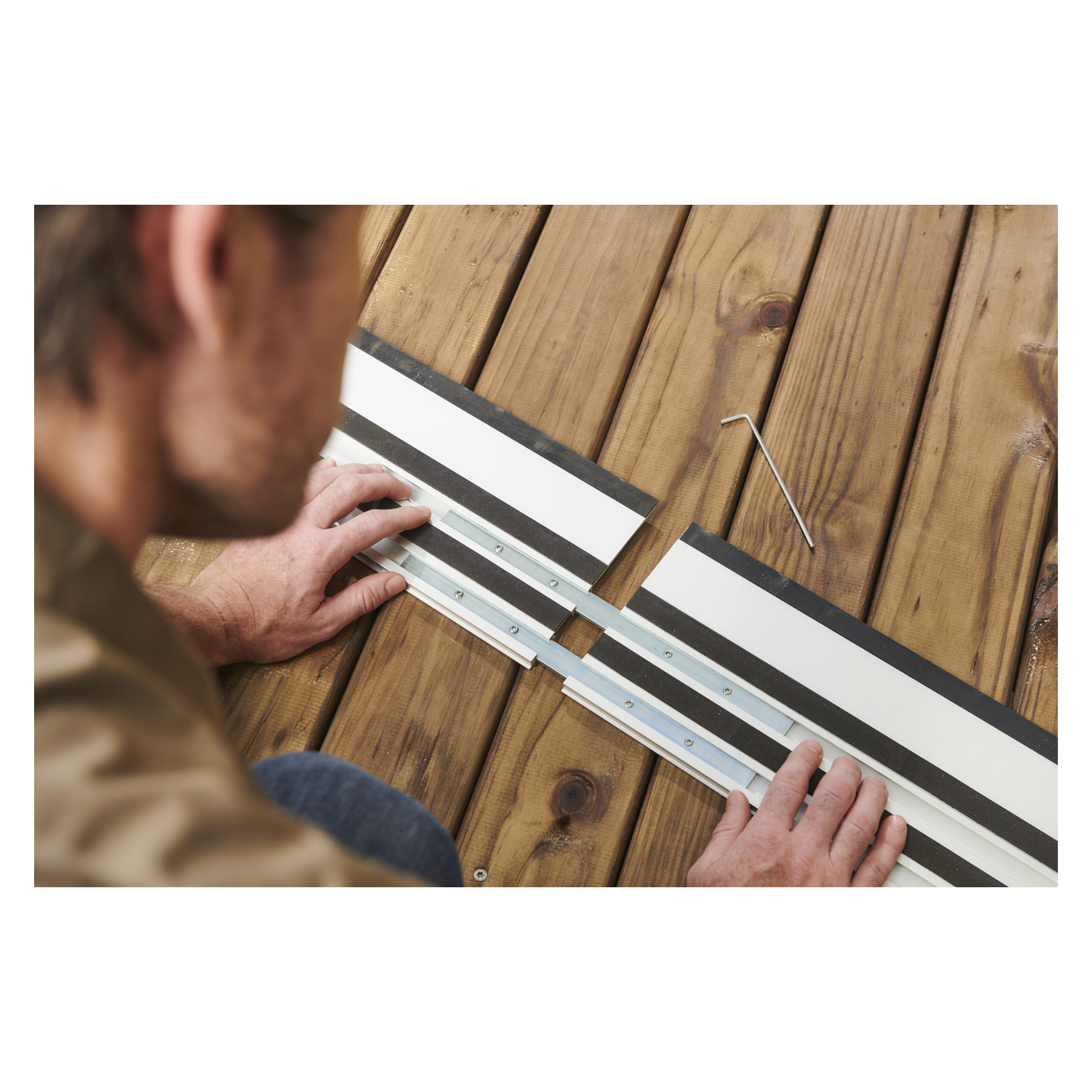 En person riktar in två Ryobi One+ HP Guide Rails för en plungesåg på trä med skruvar, en L-formad insexnyckel i närheten, ansiktet ur fokus.