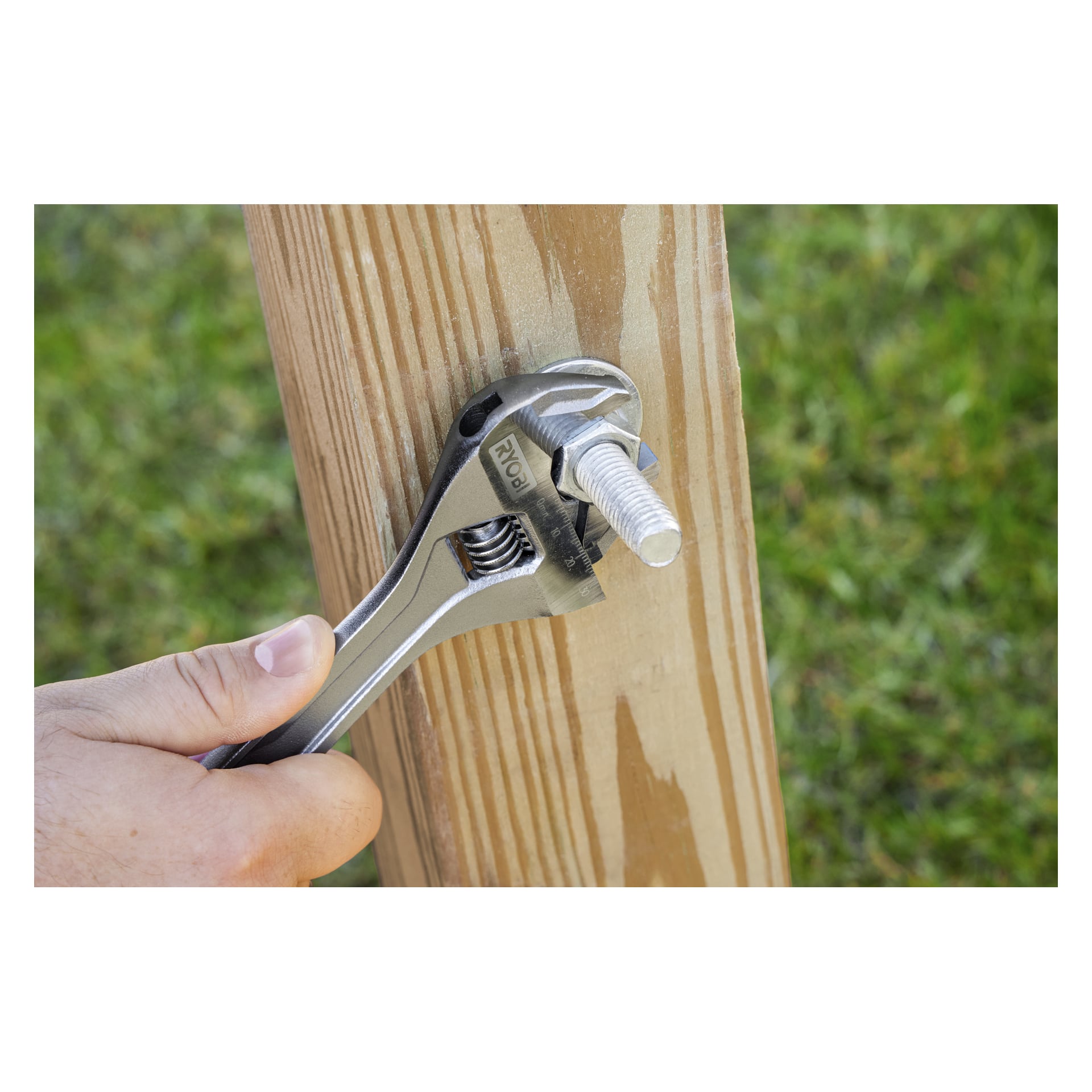 En hand drar åt en bult på en vertikal trästolpe med hjälp av Ryobi Adjustable Spanner RHAW200; gräset är i bakgrunden.