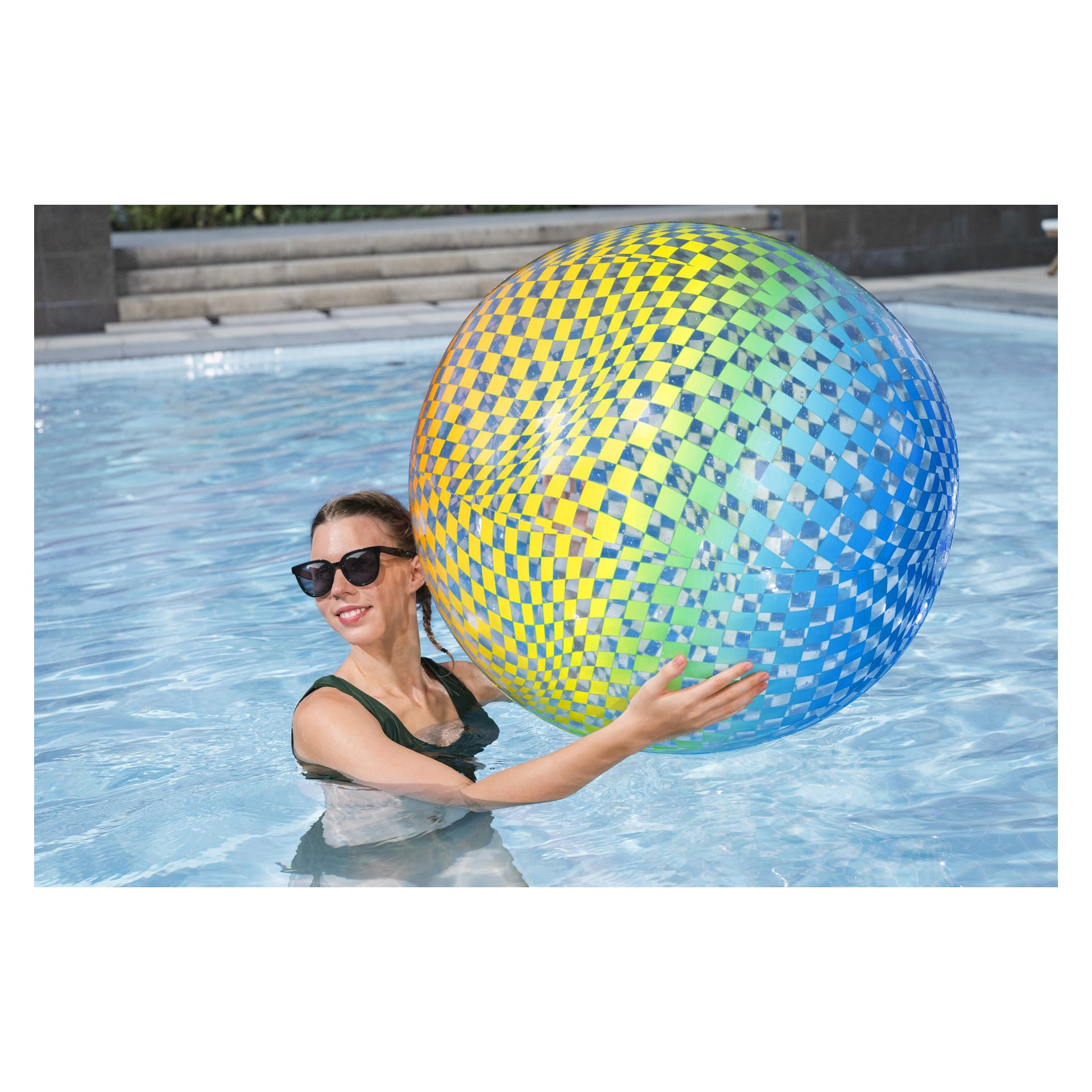 En kvinna med solglasögon och mörk baddräkt står i en pool med en Bestway Colosphere Beach Ball, trappsteg och kakel bakom sig.