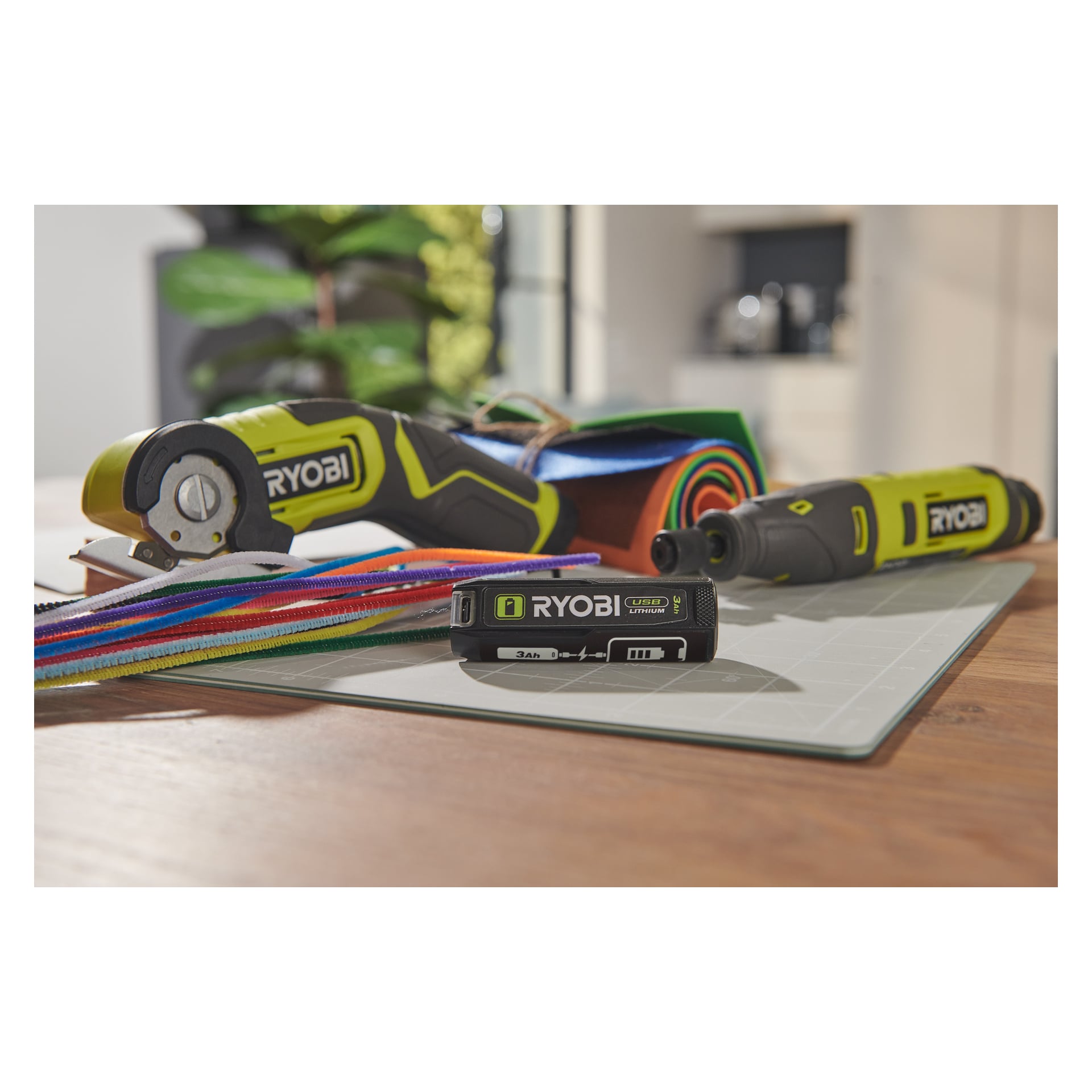 Ett par Ryobi USB litiumbatterier RB4L30X2 ligger på en skärmatta, omgiven av Ryobi-verktyg och pysselmaterial på ett träbord.