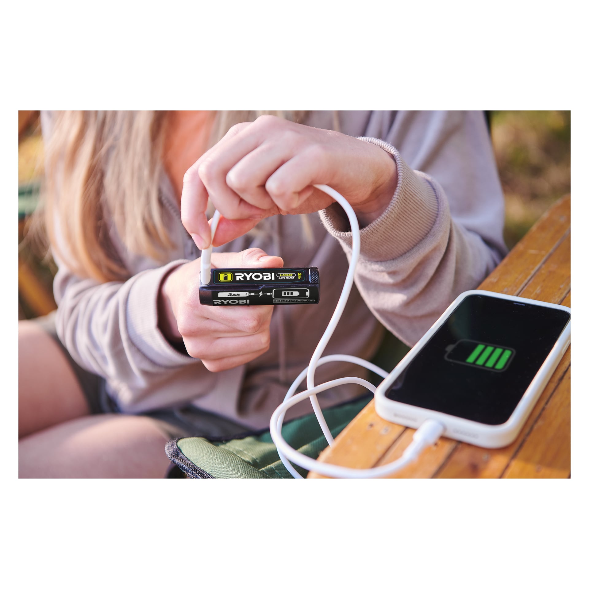 Någon kopplar en vit kabel till en svart och gul Ryobi USB Lithium Batteries RB4L30X2, laddar en telefon med en grön batteriikon.