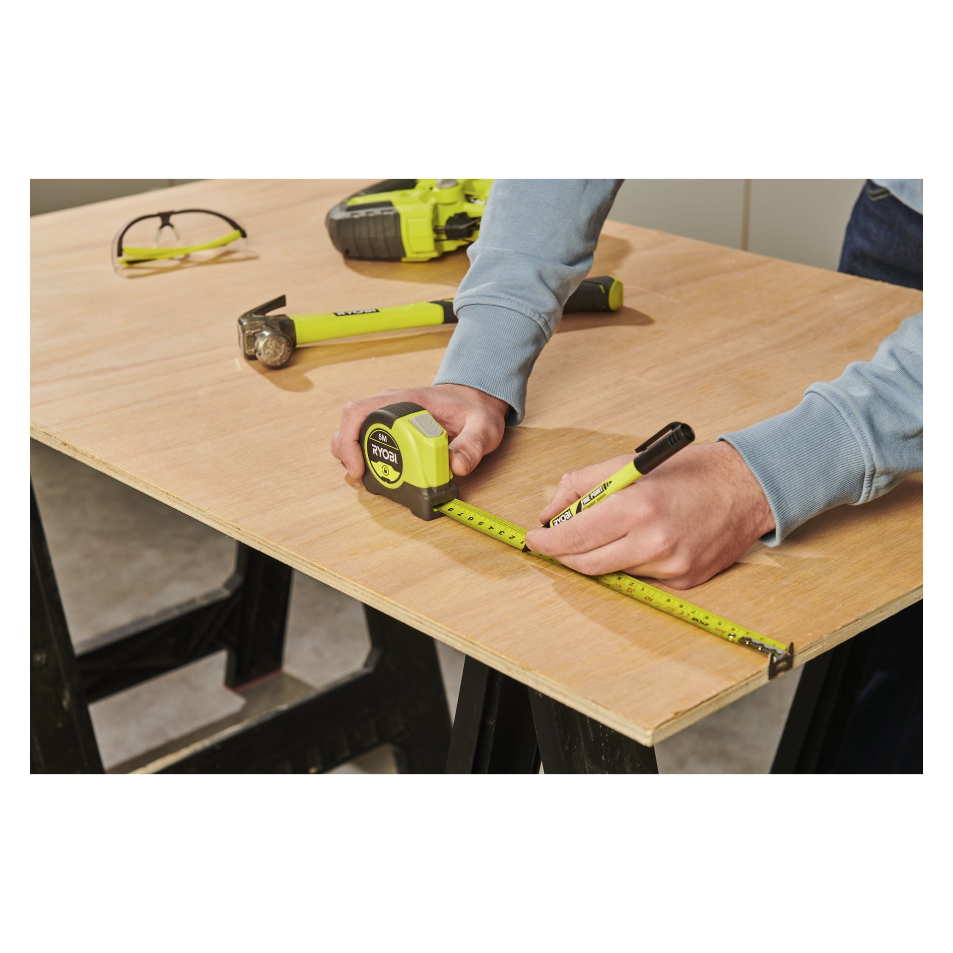 Någon mäter plywood med ett Ryobi-måttband, RHMT5 och marker; En borrmaskin, skyddsglasögon och en hammare ligger i närheten på bordet.