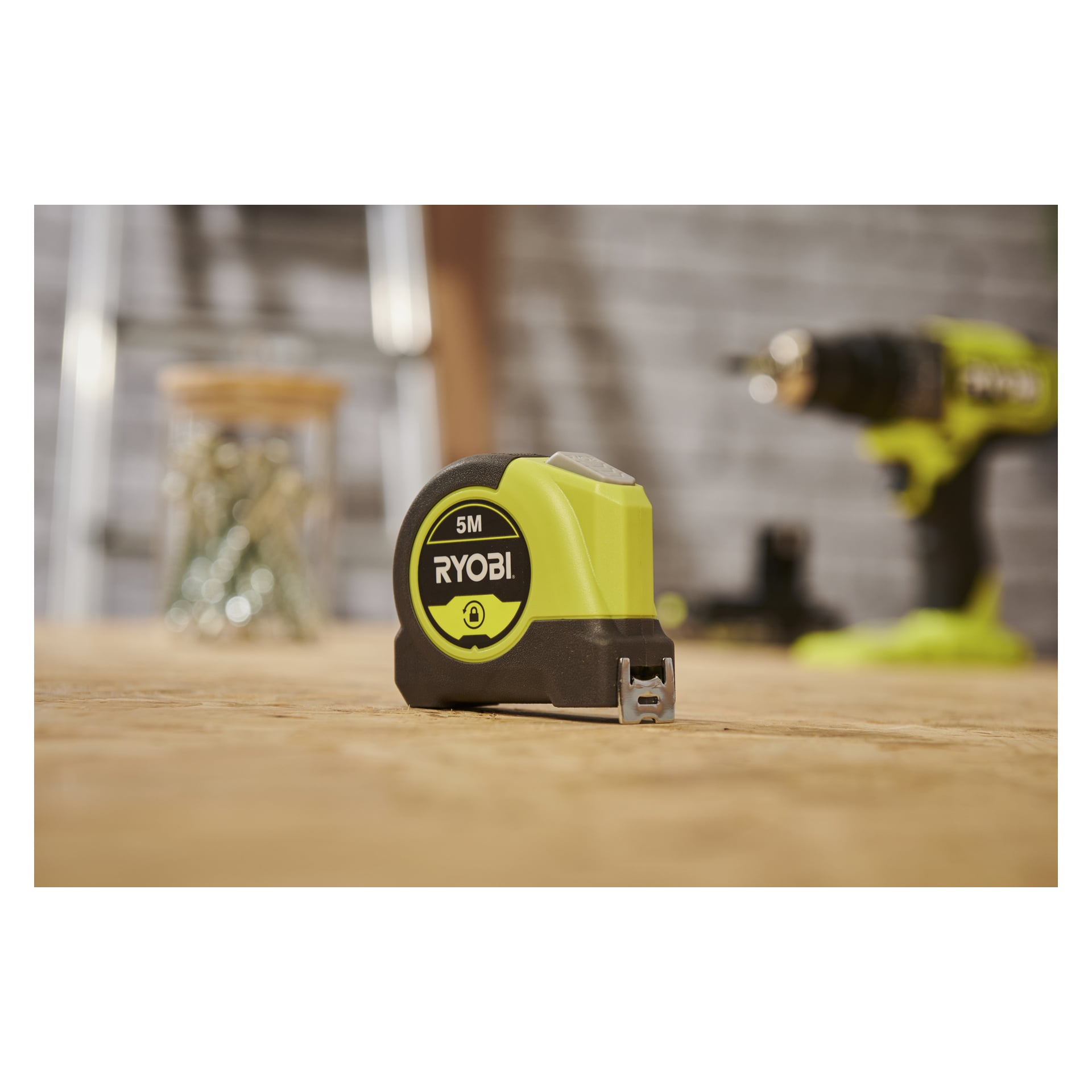 Ett Ryobi RHMT5-måttband står på trä, med en suddig borr och en skruvburk i bakgrunden, vilket påminner om en verkstadsmiljö.