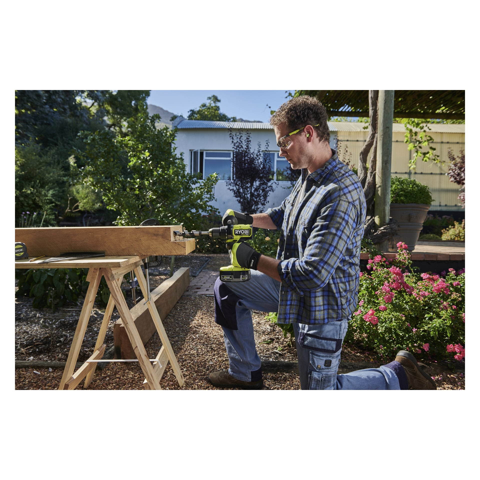 Iført arbeidsklær bruker en mann en Ryobi One+ HP-skrutrekker for å sette sammen tre på sagbukker i en hage ved et hus.