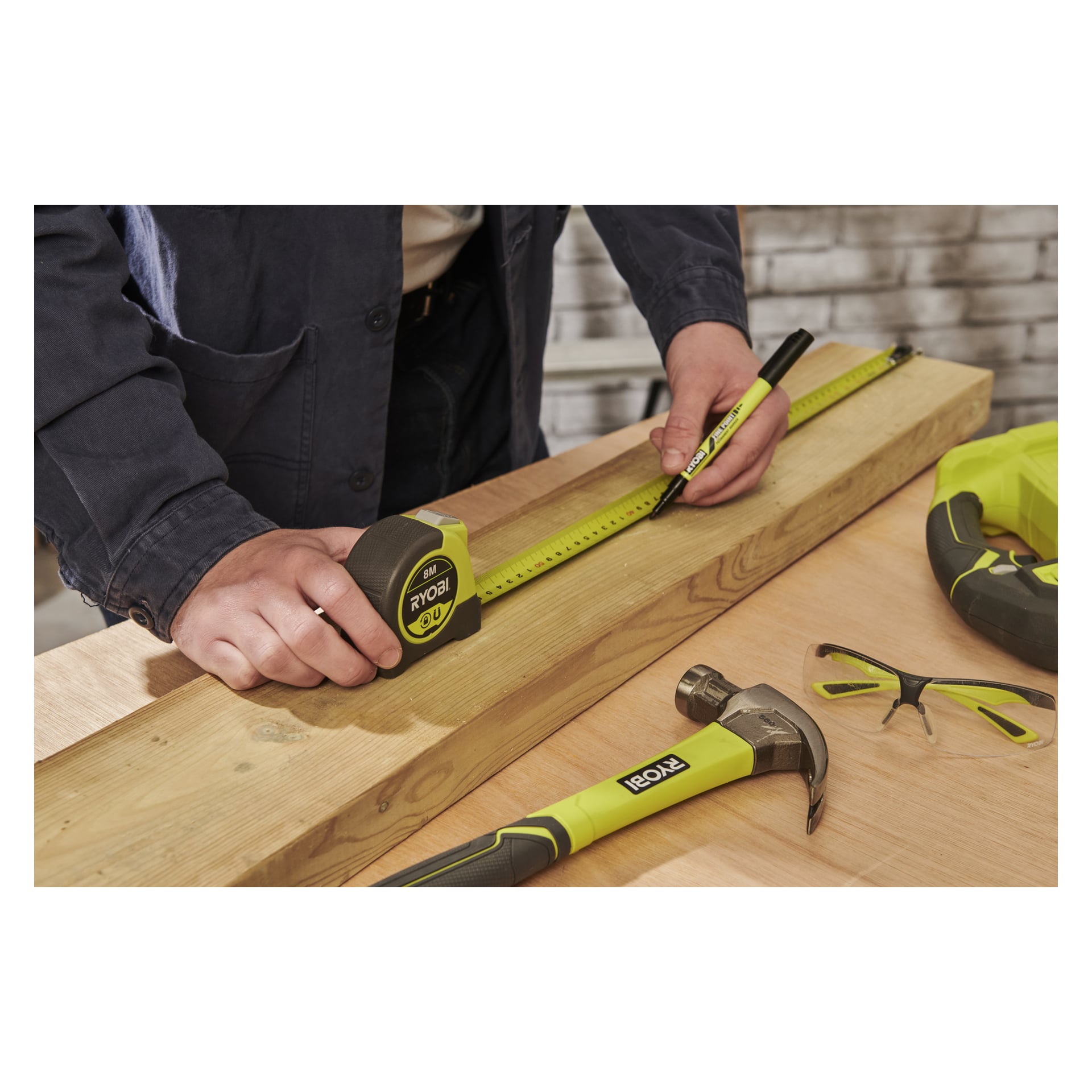 Någon mäter en träplanka på en arbetsbänk med ett Ryobi-måttband och en markör; i närheten finns en Ryobi-hammare, skyddsglasögon och ett elverktyg.