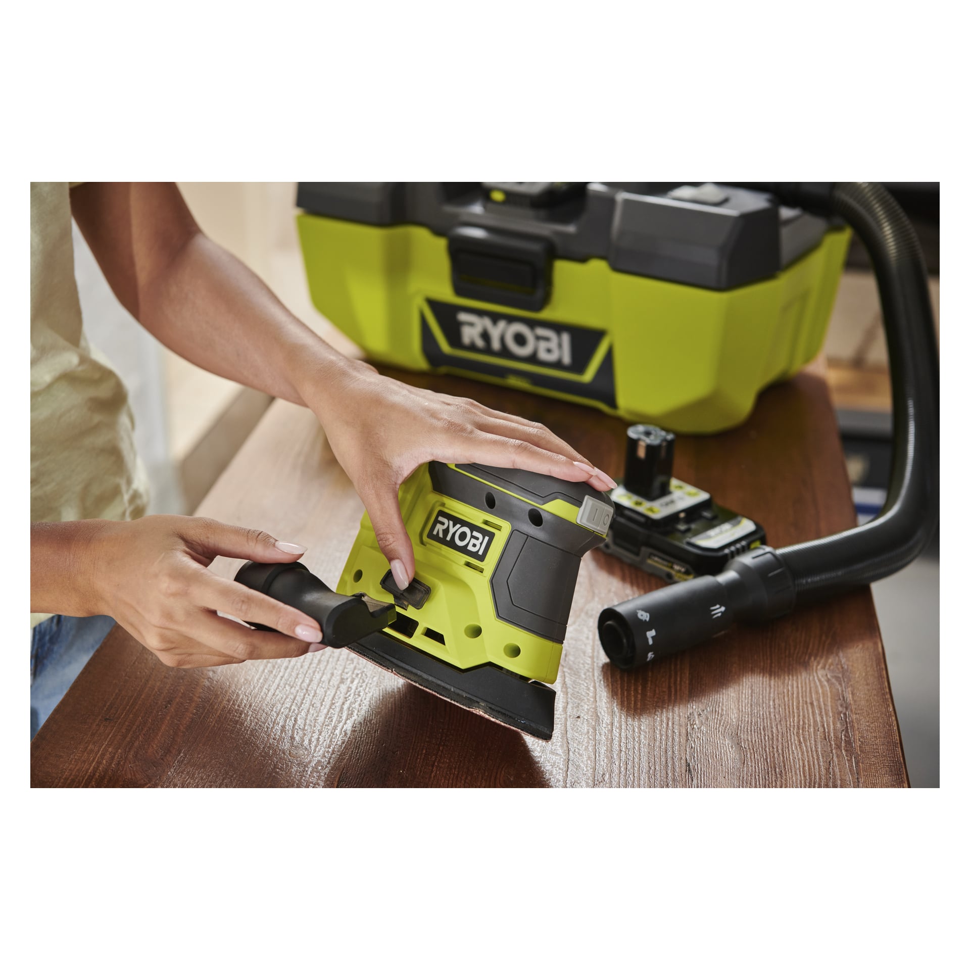 Någon dammsuger damm från en Ryobi One+ Multi-slipare R18PSN-0 på träbrädor, med ett Ryobi-fodral och verktyg synliga bakom.