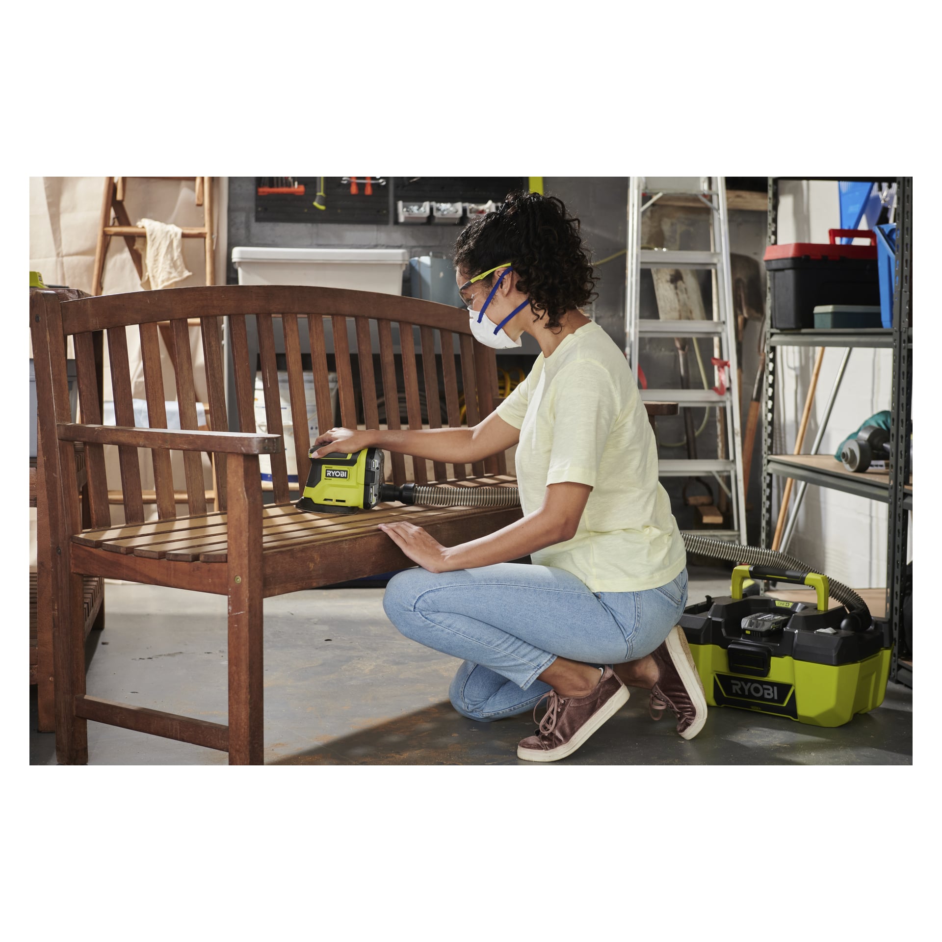 En kvinna i skyddsutrustning slipar en träbänk med en Ryobi One+ Multi-slipare R18PSN-0 kopplad till en dammsugare.