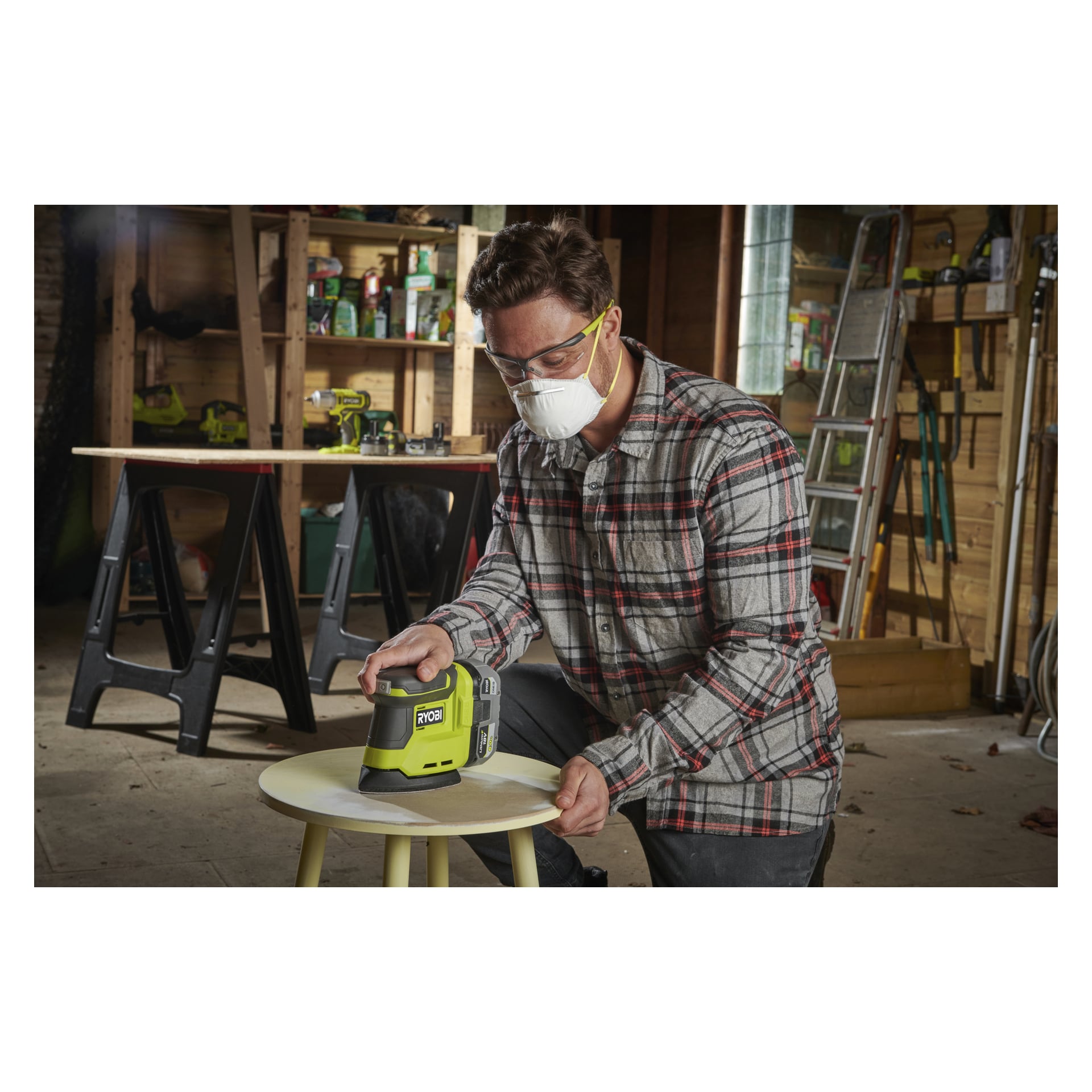En person med skyddsglasögon, ansiktsmask och rutig skjorta slipar en rund pall med en Ryobi One+ slipmaskin i en verkstad fylld med verktyg.
