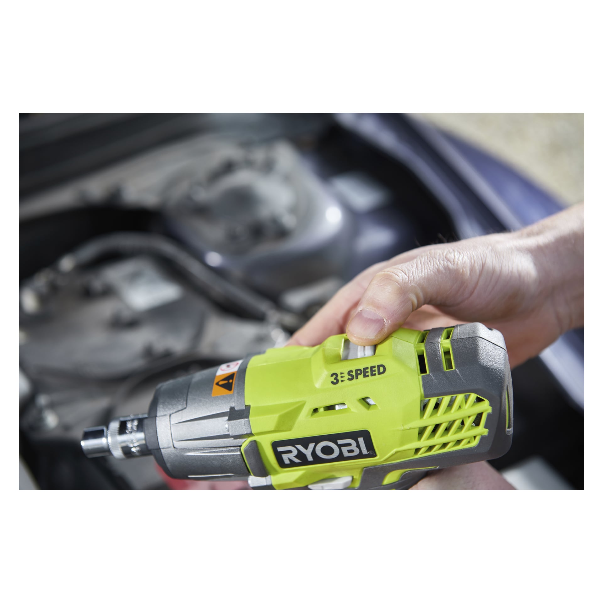 En persons hand greppar en grön och silverfärgad Ryobi One+ slagnyckel RIW183N-0, med synlig brandmärkning, framför en suddig bilmotor.