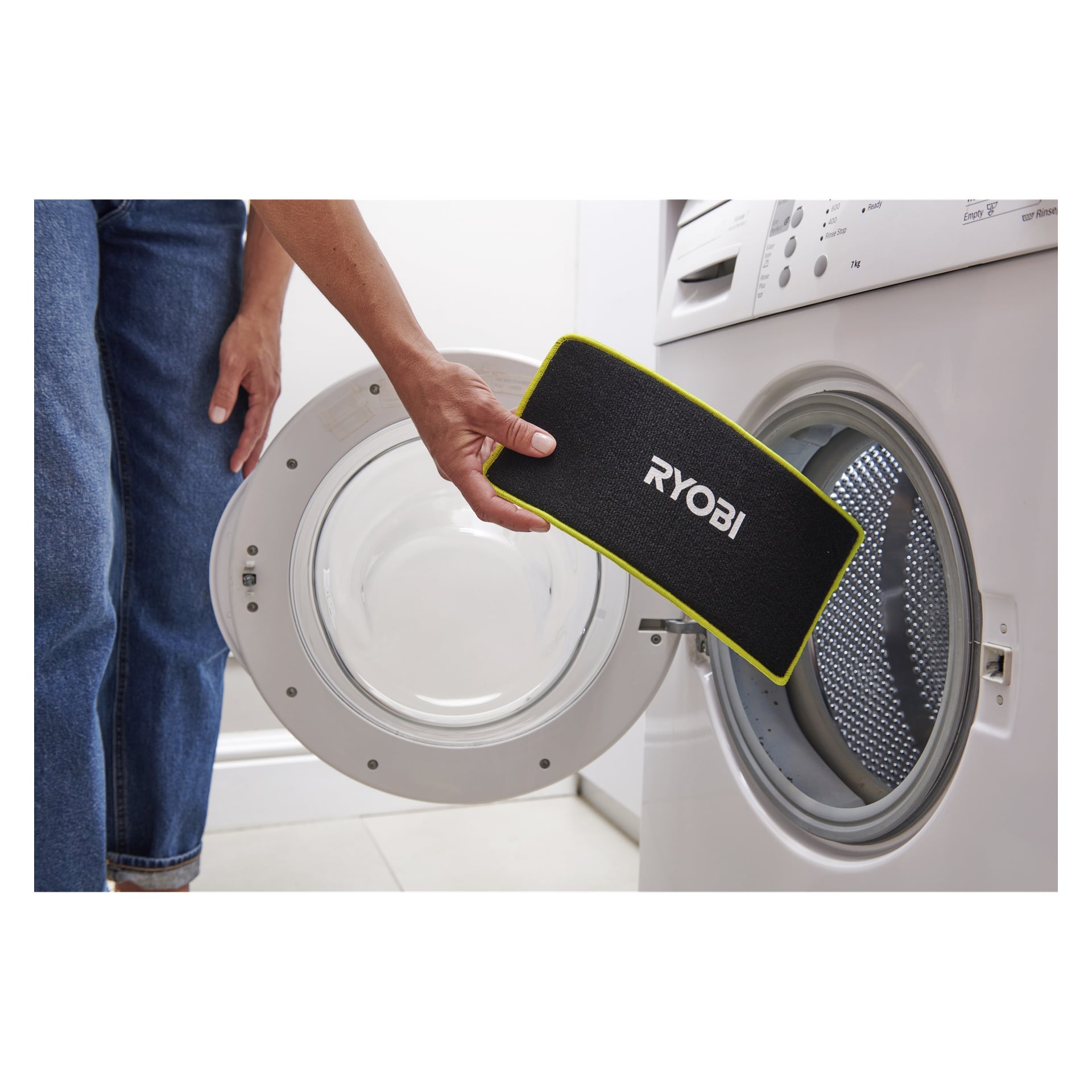 En person i blå jeans och vit skjorta håller i en svart Ryobi USB Lithium Mopp-tygpåse nära en öppen frontmatad tvättmaskin.