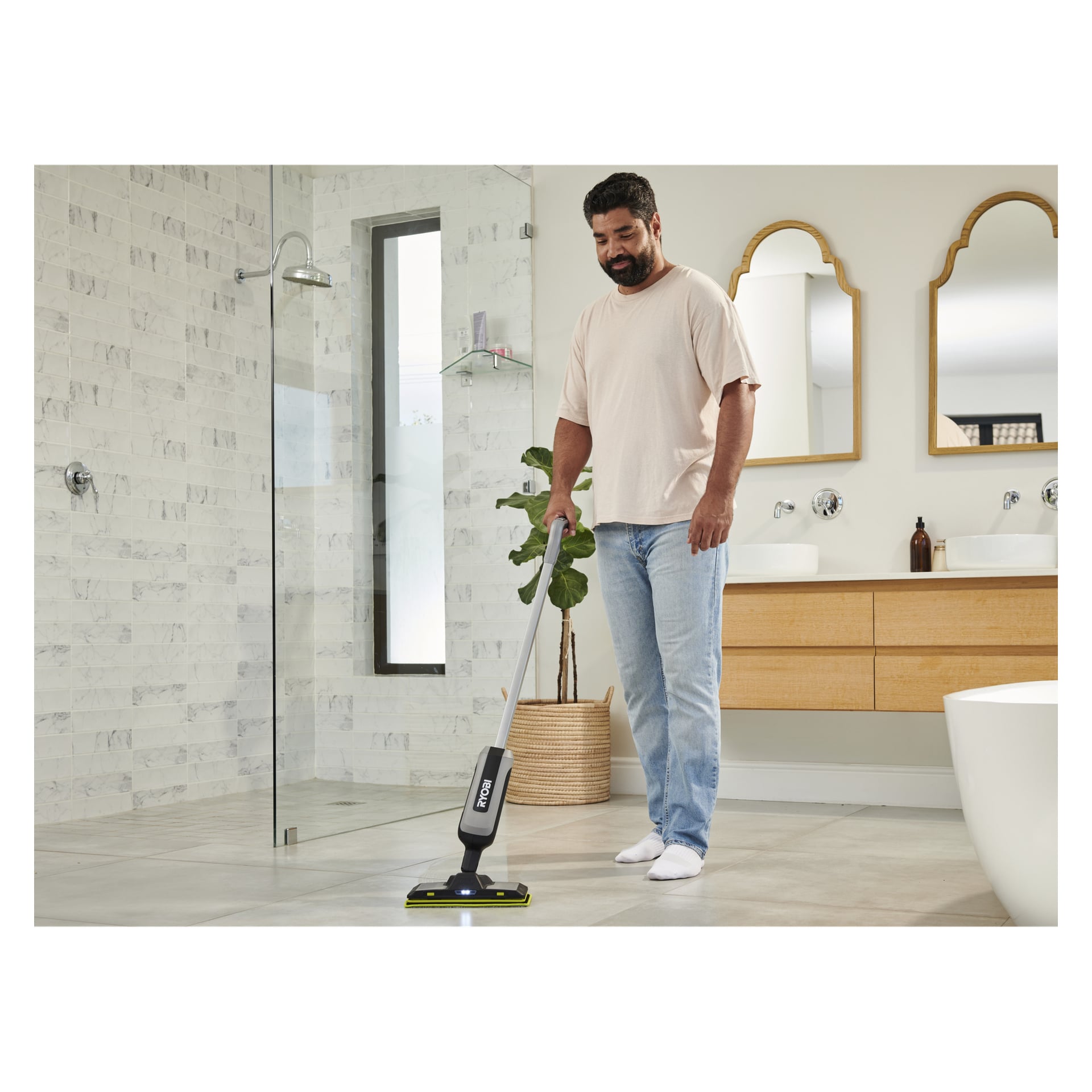 En man i en tunn t-shirt och jeans rengör ett kaklat modernt badrumsgolv med Ryobi USB Lithium Mop-tyg RSM4A1 3-pack.