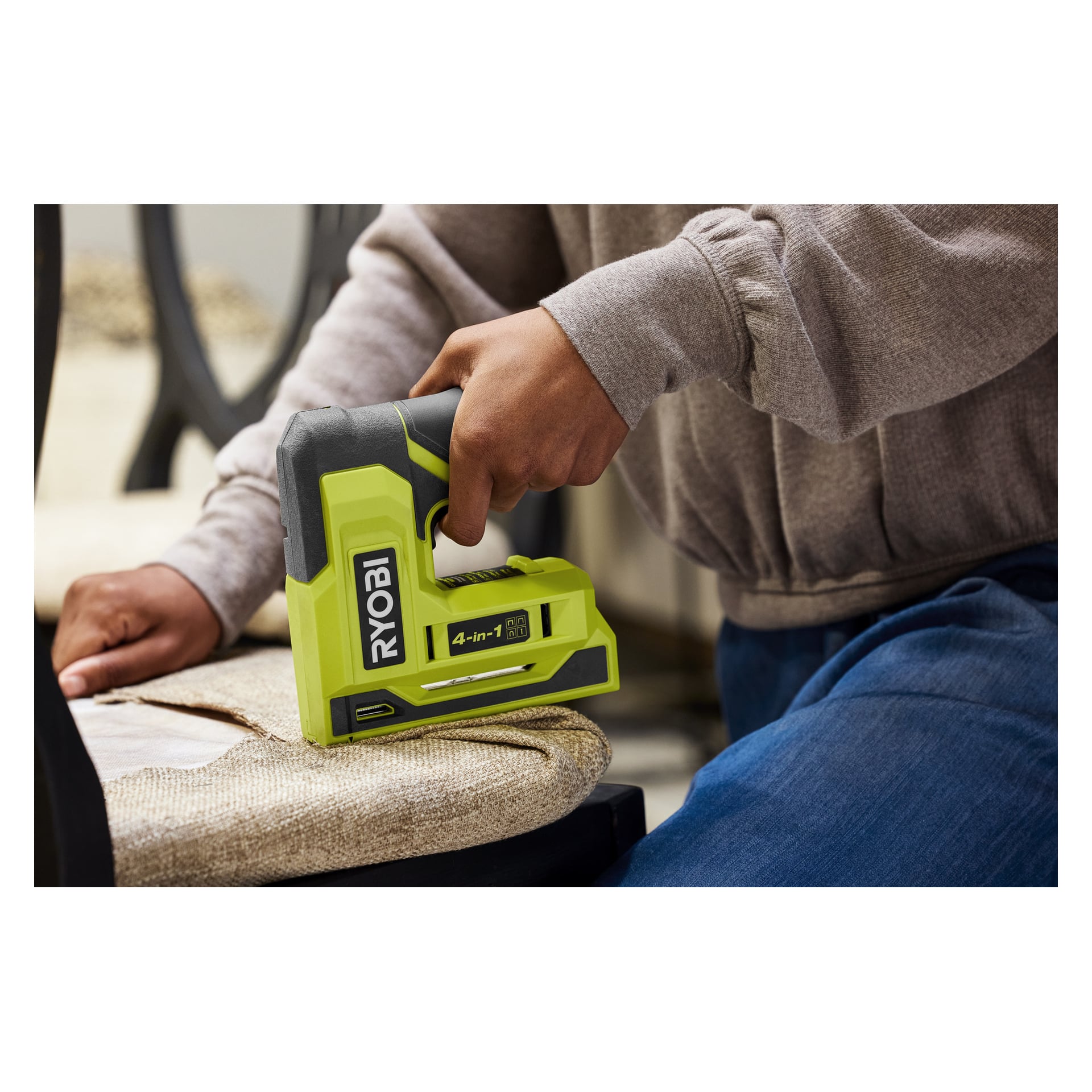 En person i en beige genser og blå jeans bruker en grønn Ryobi-stiftepistol for å feste blekt stoff på en trestolramme.