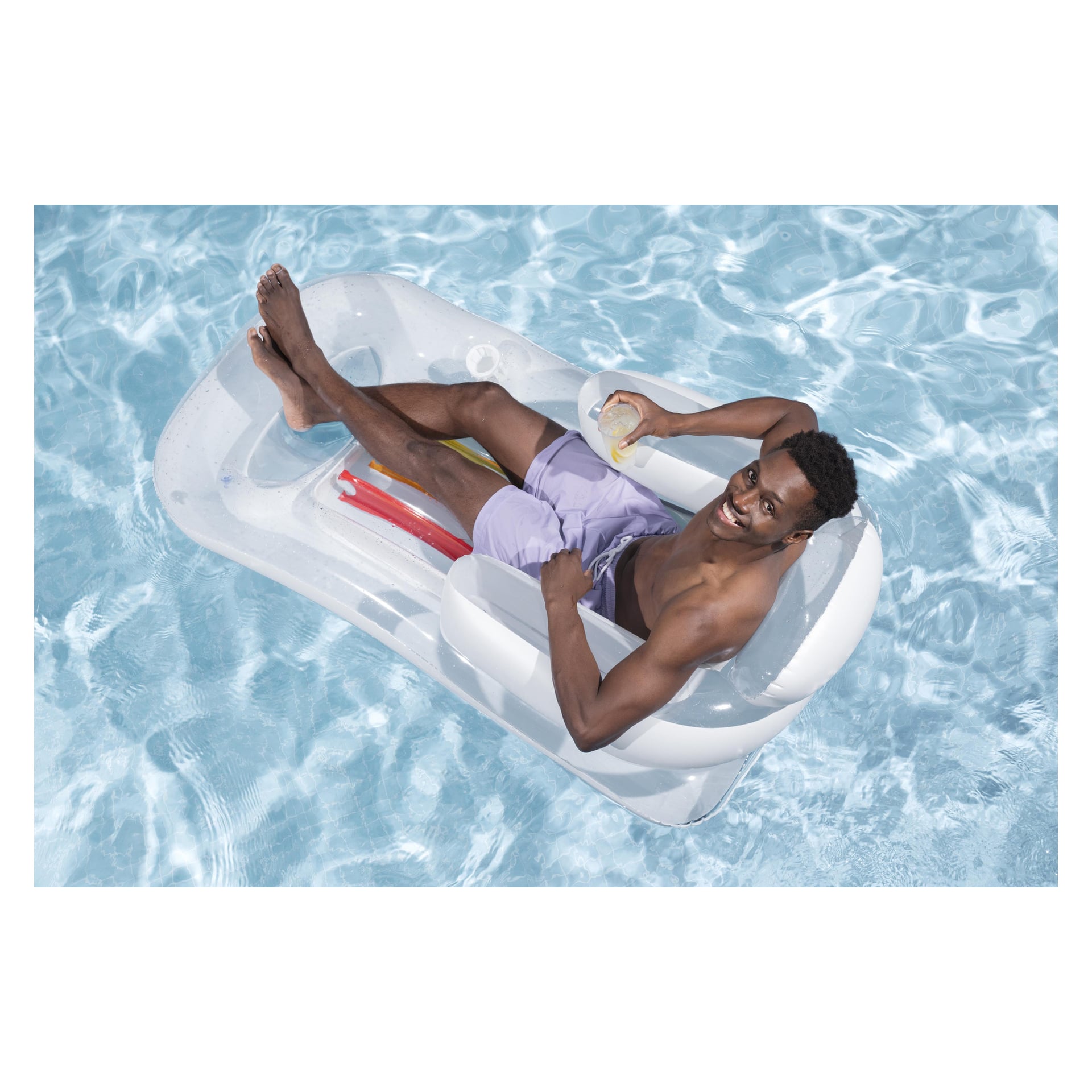En man slappnar av på en färgglad Bestway Sun-madrass, flytande i en klar pool, iklädd lila badbyxor och ler med en drink i handen.