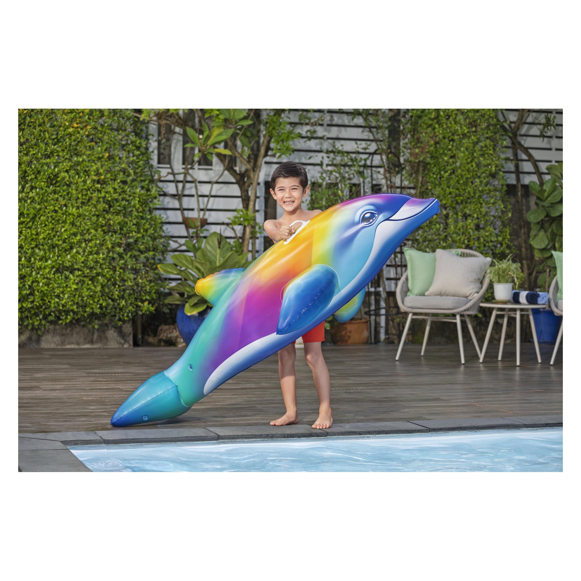 Wesoły chłopiec w czerwonych szortach stoi przy basenie z Bestway Dolphin Daydream Ride-On, z zielonymi roślinami i krzesłami na zewnątrz za sobą.