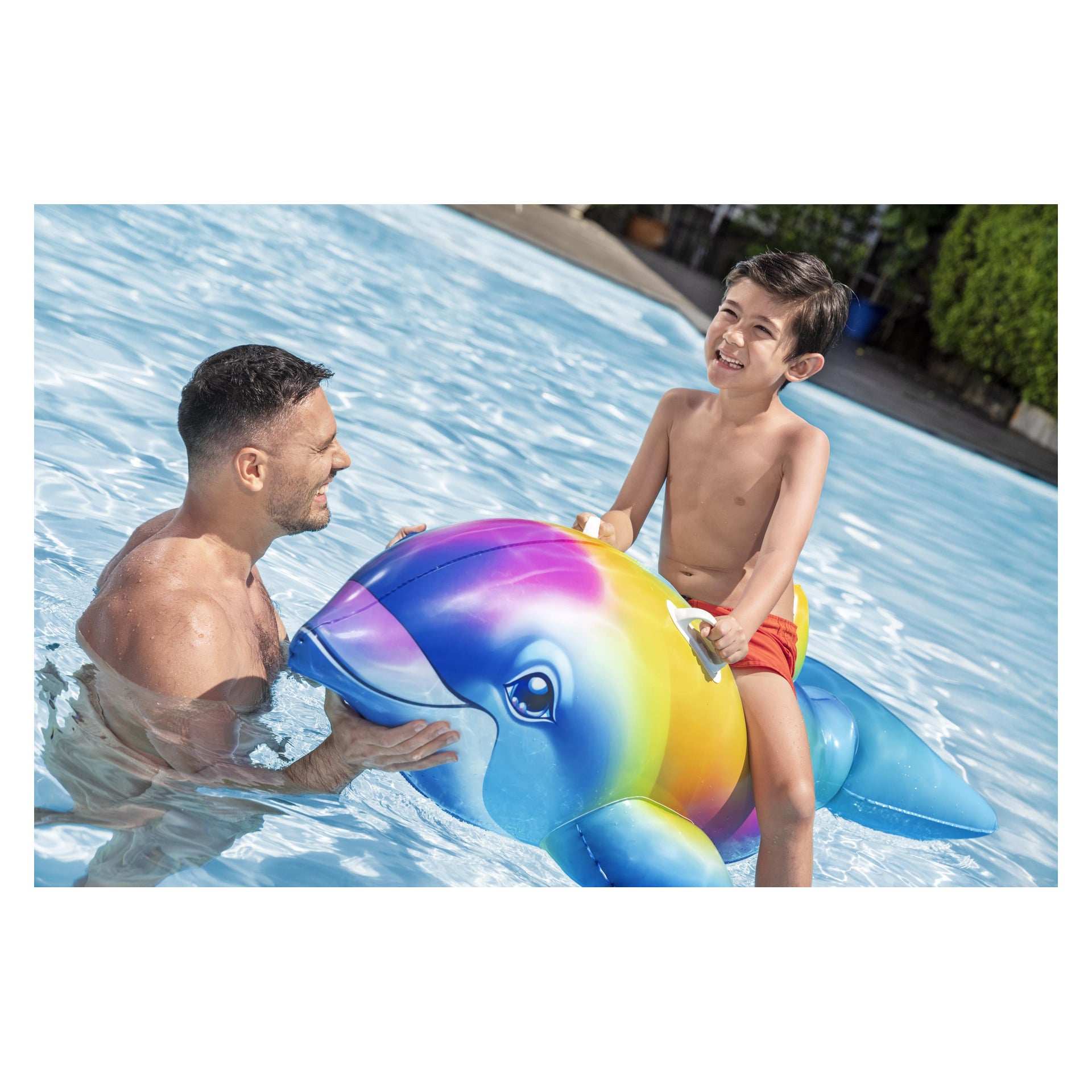 Dziecko w czerwonych kąpielówkach siedzi na kolorowym Bestway Dolphin Daydream Ride-On w basenie, a dorosły stabilizuje platformę.