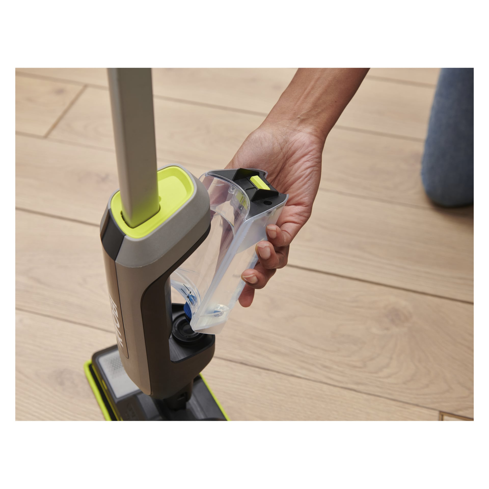 Någon kopplar loss den klara vattentanken från en Ryobi USB Lithium spraymopp med grått och grönt handtag på ett ljust trägolv.