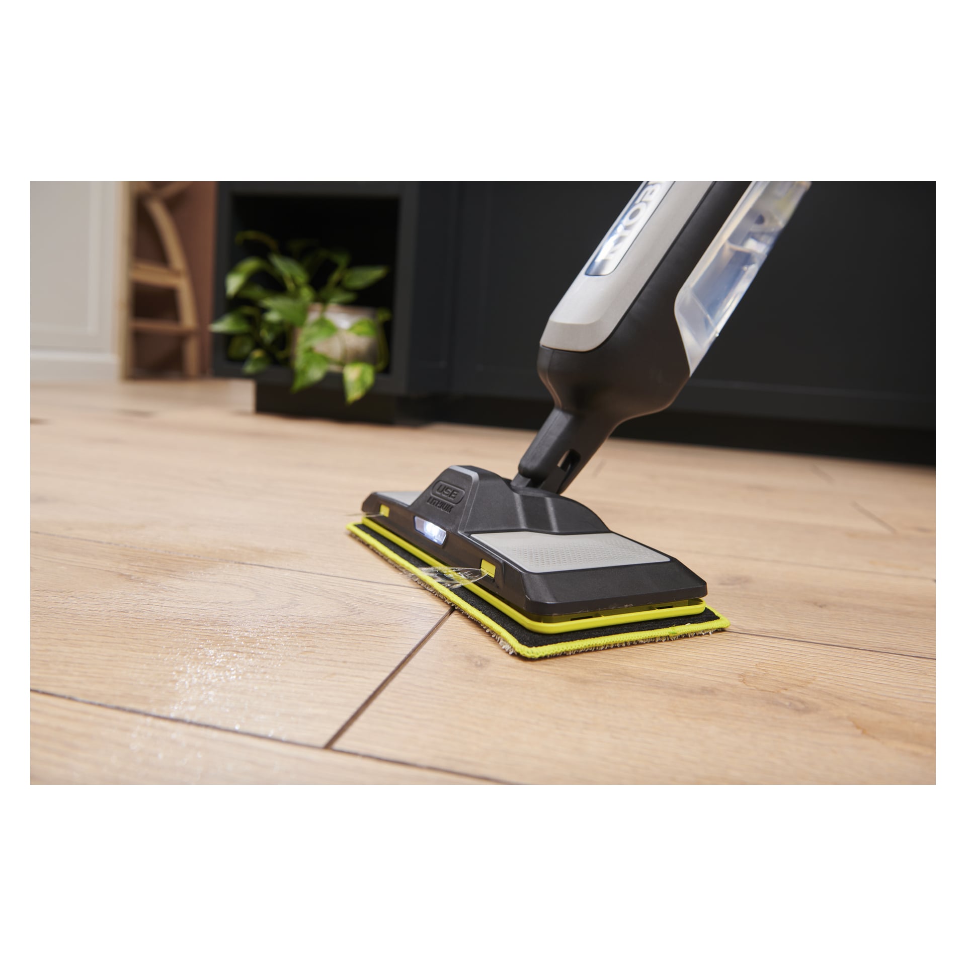 Närbild av Ryobi USB Lithium Battery Driven Spray Mop RSM4-120G som rengör ett lätt trägolv, med en växt och en stol i närheten.
