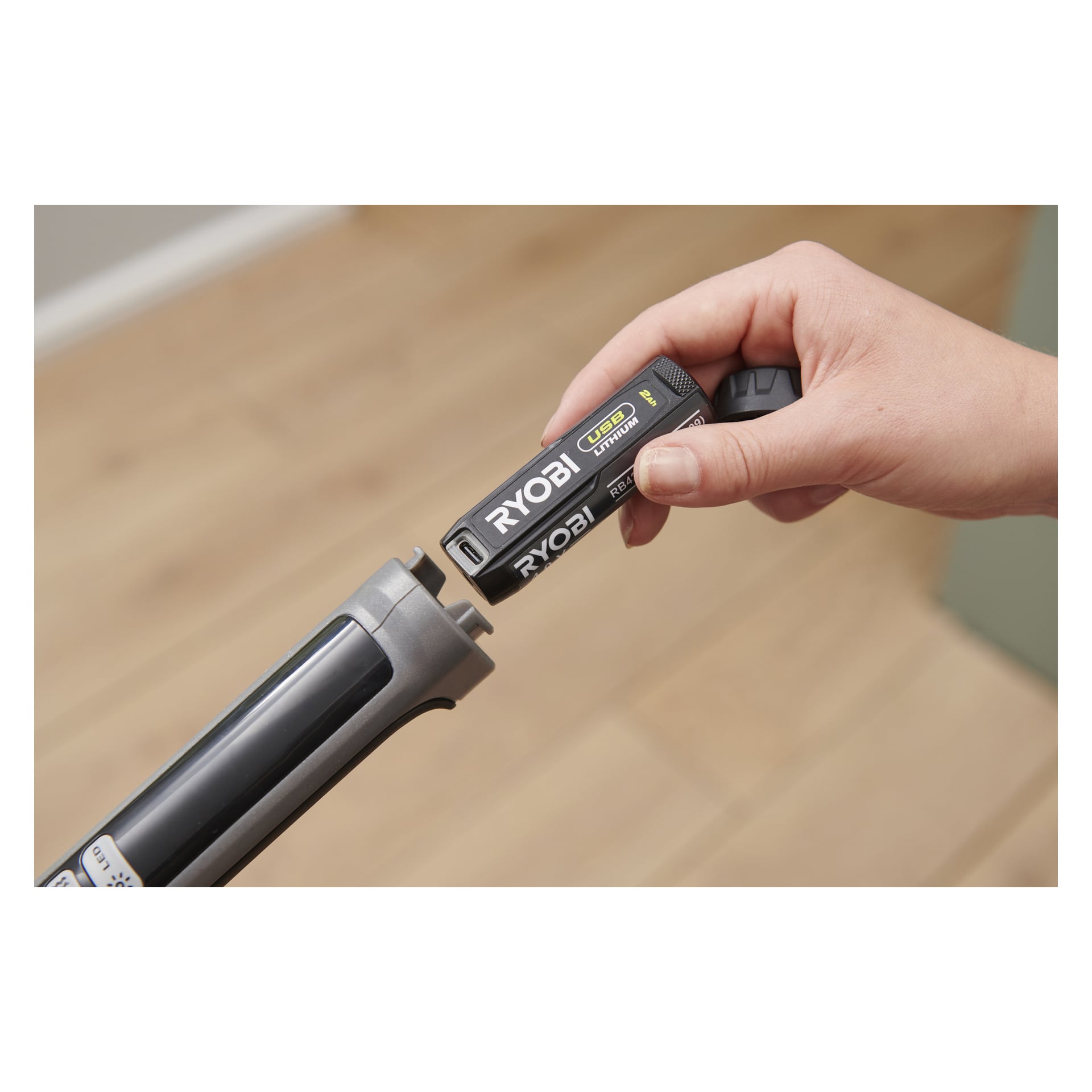 En hand placerar ett Ryobi USB Lithium-batteri i den grå RSM4-120G spraymoppen, med ett suddigt trägolv i bakgrunden.