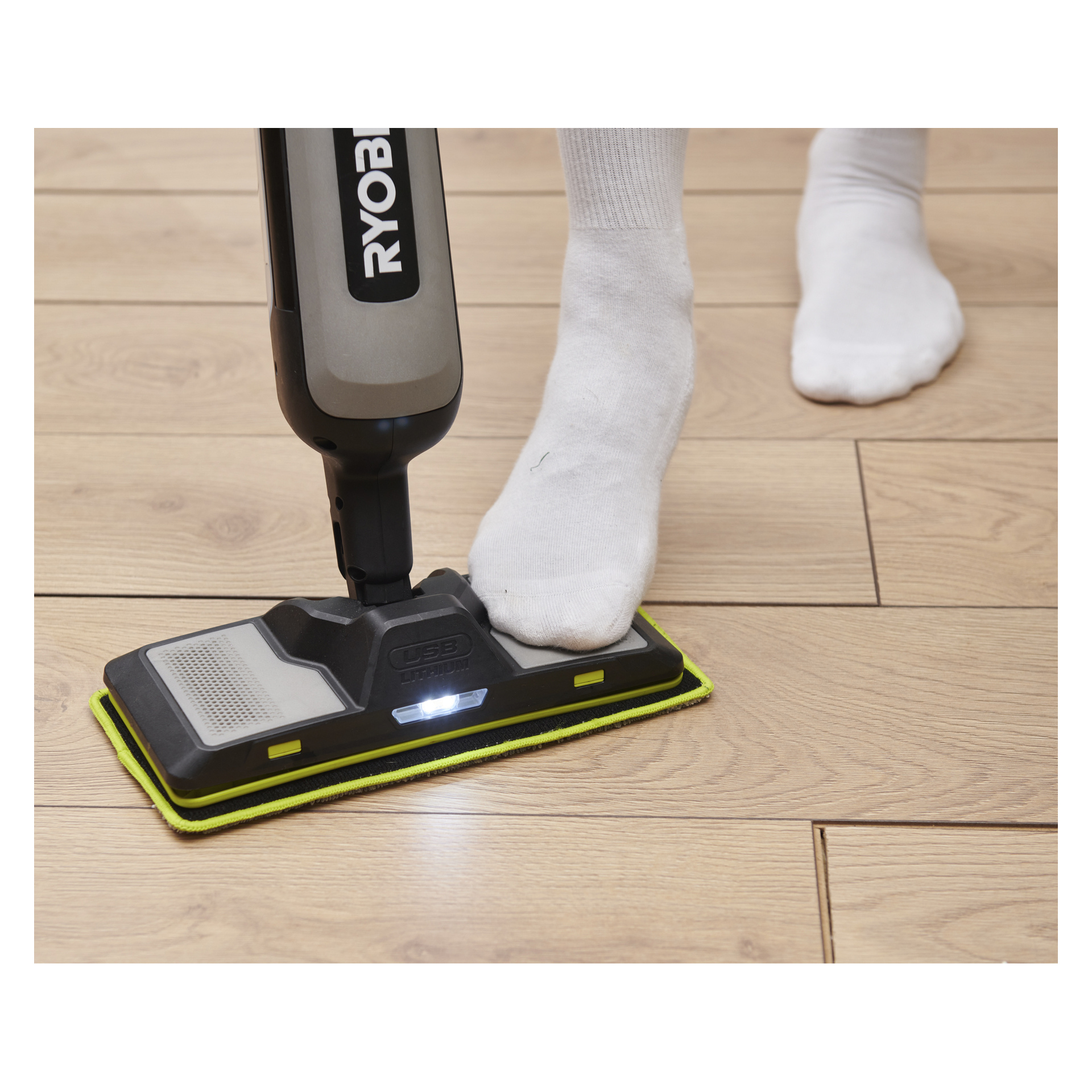 En person i vita strumpor använder sin fot för att styra en Ryobi RSM4-120G spraymopp med LED-lampa på ett lätt trägolv.