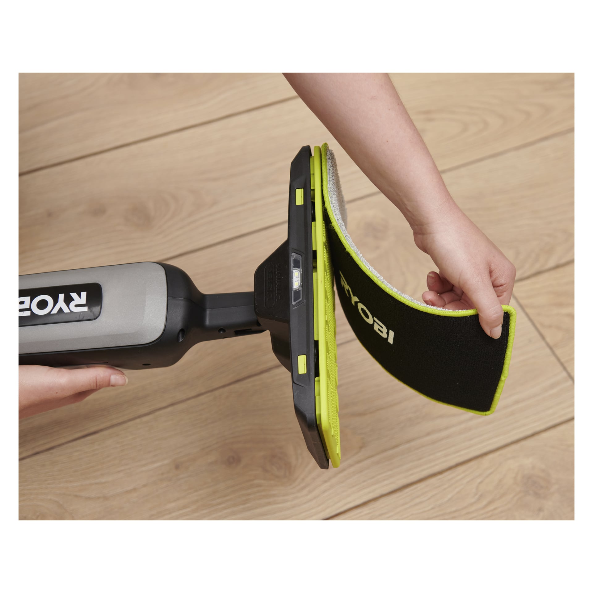 En person fäster eller lossar en rektangulär platta från Ryobi USB Lithium Spray Mop RSM4-120G på ett trägolv.