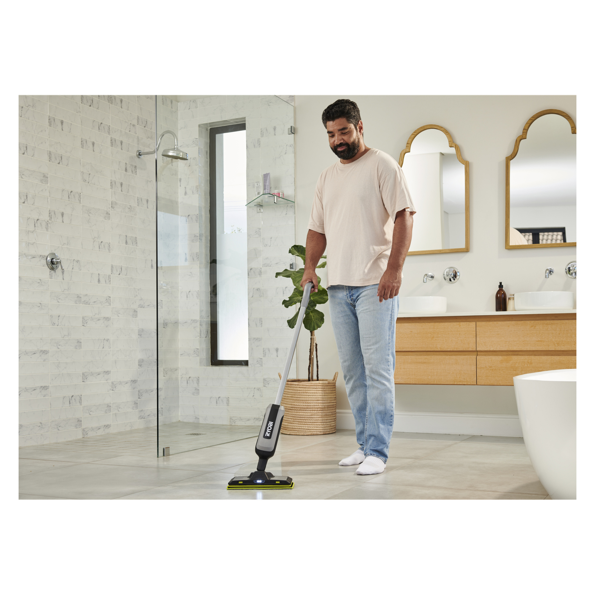 En man i beige t-shirt och jeans rengör ett modernt badrumsgolv med en Ryobi-spraymopp, omgiven av växter och elegant inredning.