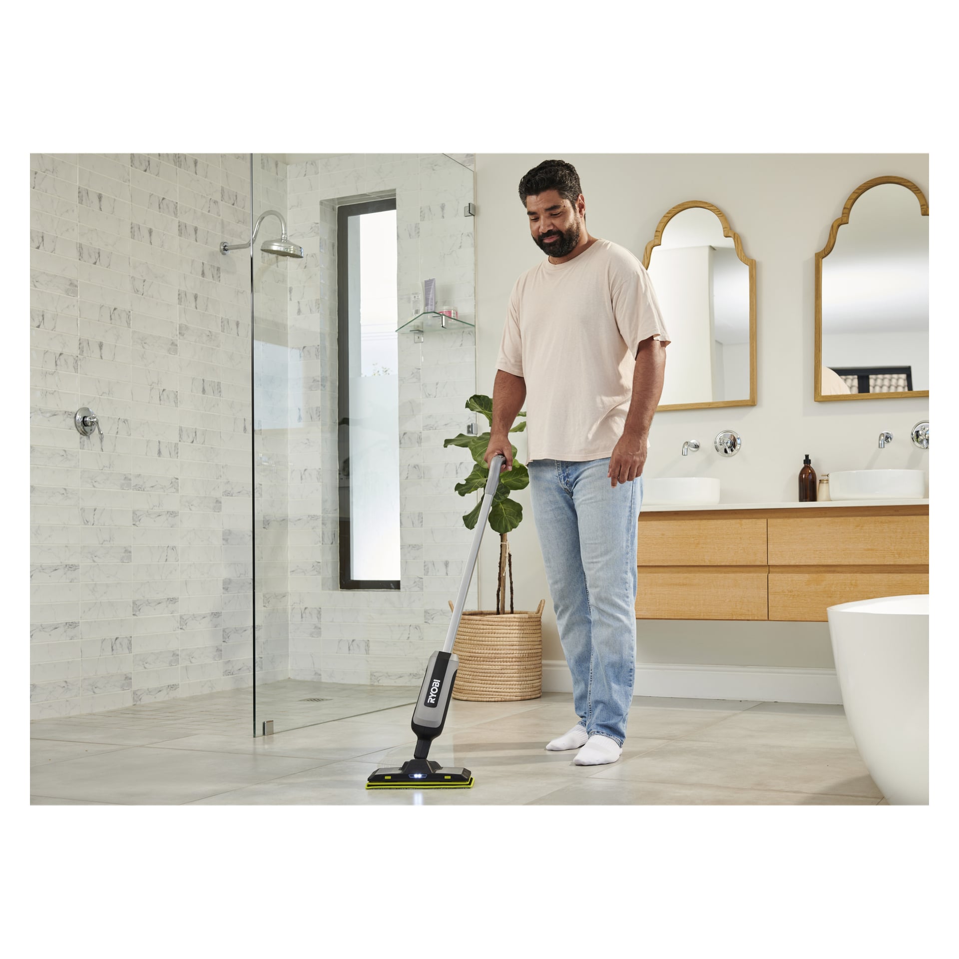 En man i beige t-shirt och jeans rengör ett modernt badrumsgolv med en Ryobi-spraymopp, omgiven av växter och elegant inredning.