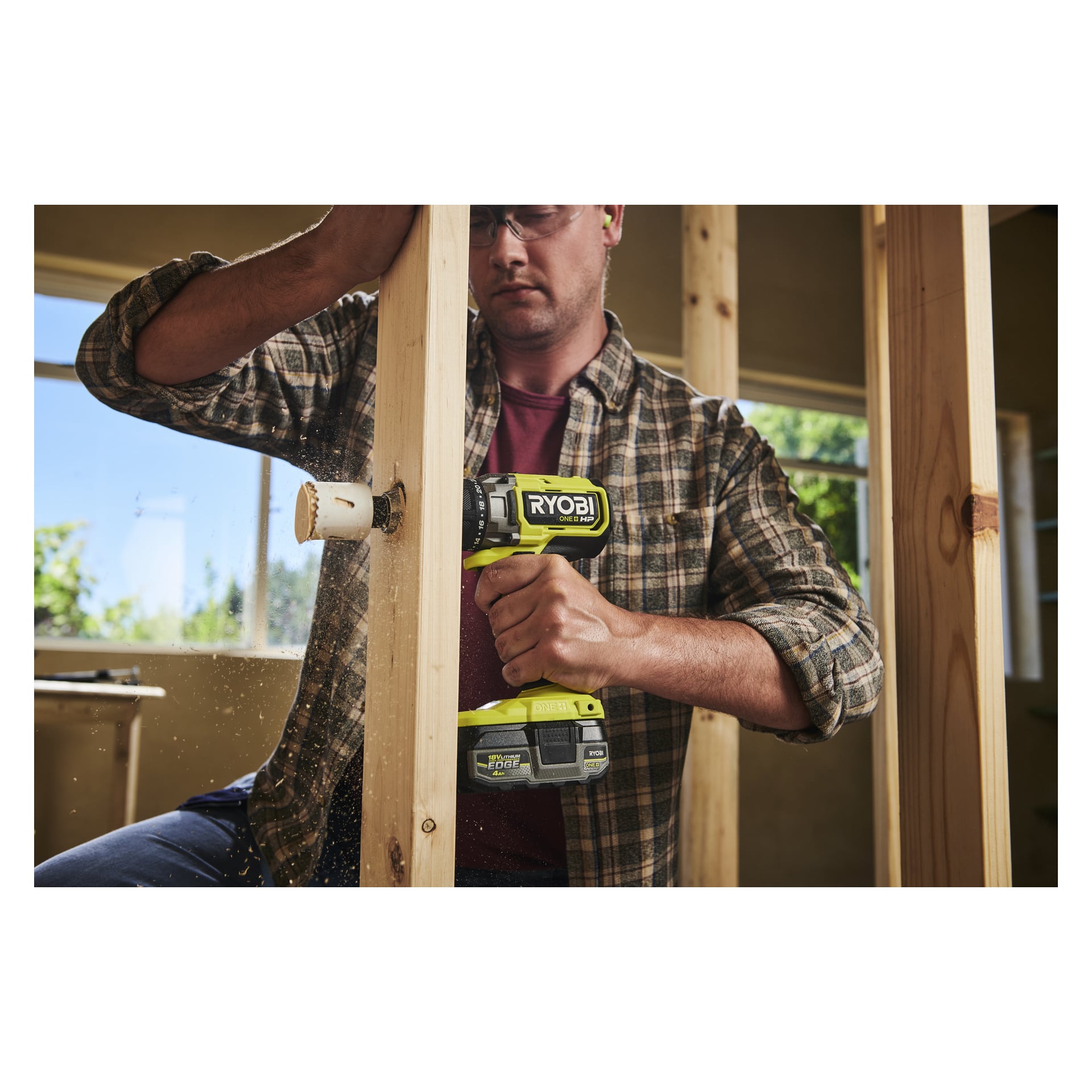 En person med briller og rutete skjorte borer inn i en trebjelke med en gul Ryobi One+ HP-skrutrekker i et uferdig rom.