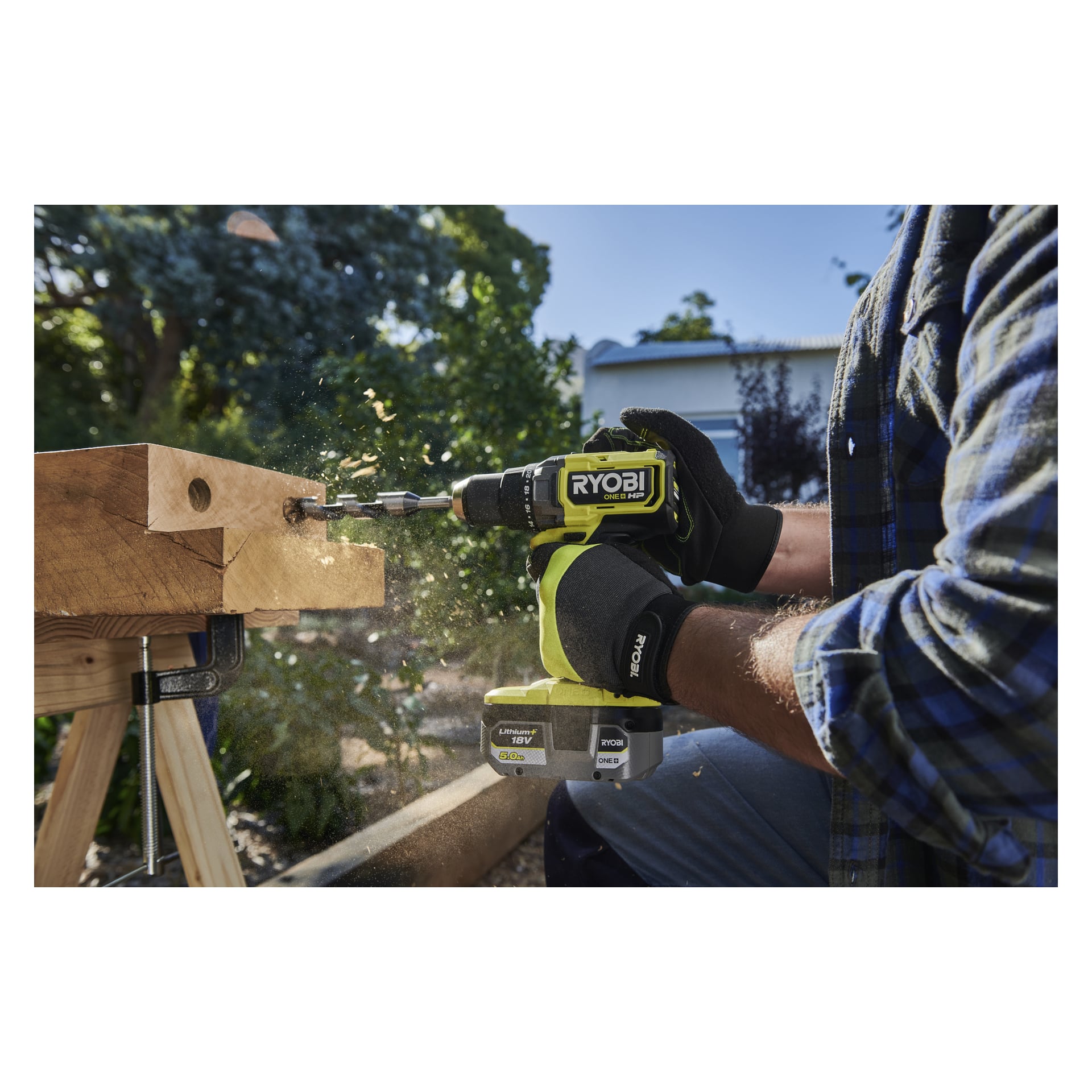 Hanskekledde hender betjener en Ryobi One+ HP skrutrekker RDD18X1-0, som borer treverk utendørs med flak som flyr, grøntområder og hus bak.