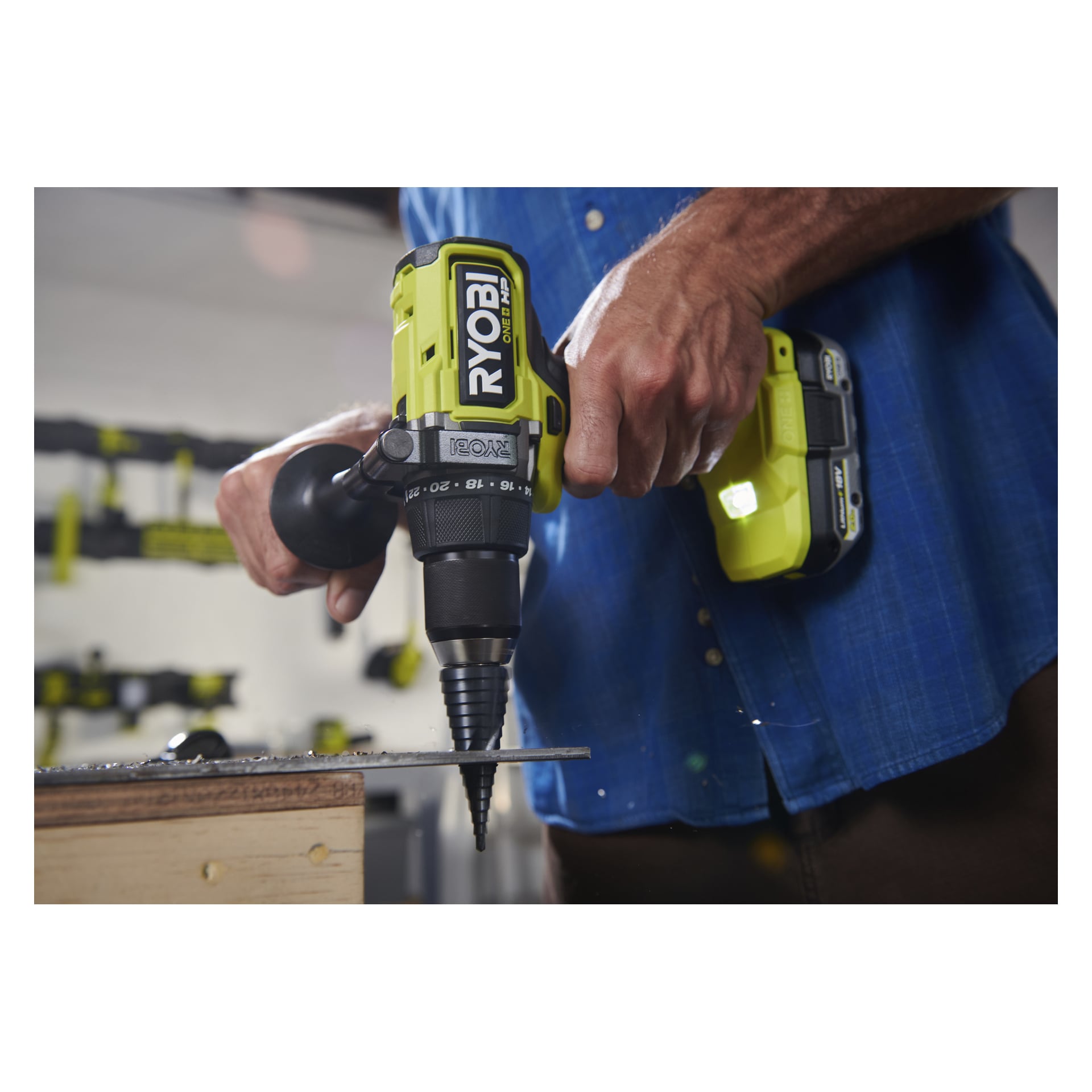 Iført en blå skjorte borer noen inn i metall med en Ryobi One+ HP RDD18X1-0; Hånden og boret deres er skarpe, bakgrunnsverktøyene uklare.