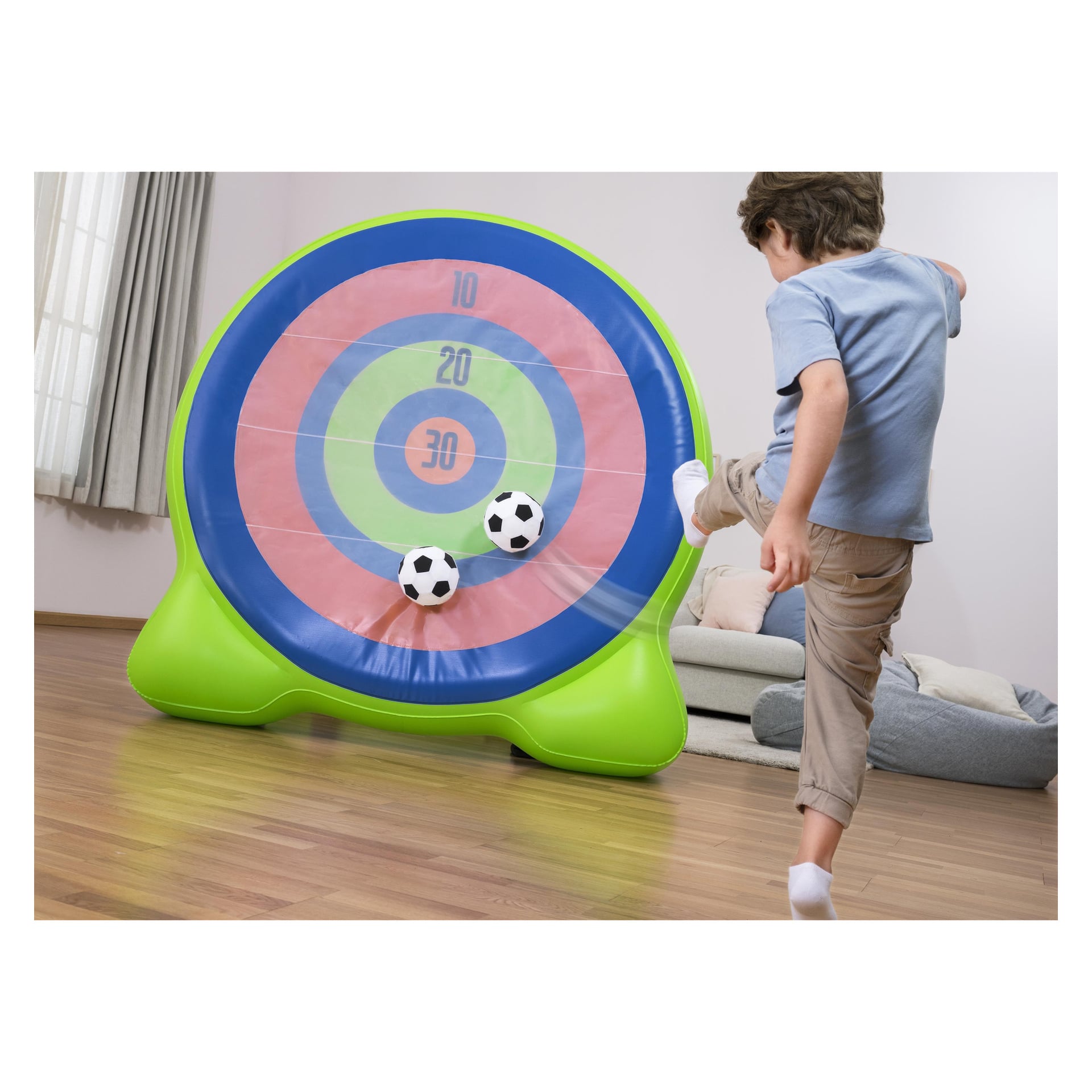 Ett barn sparkar en boll mot ett uppblåsbart dartbrädespel i ett vardagsrum med trägolv; En annan boll fastnar på målet.