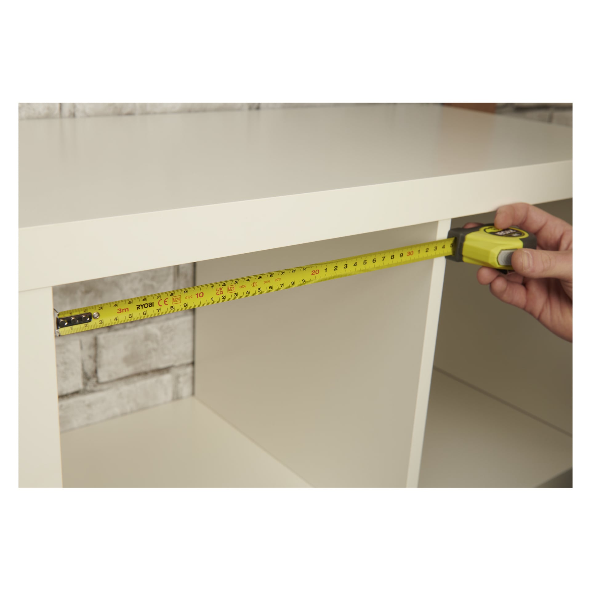 En person använder ett Ryobi Tape Measure RHMT3 för att mäta insidan av bredden på en vit hylla framför en vit tegelvägg.
