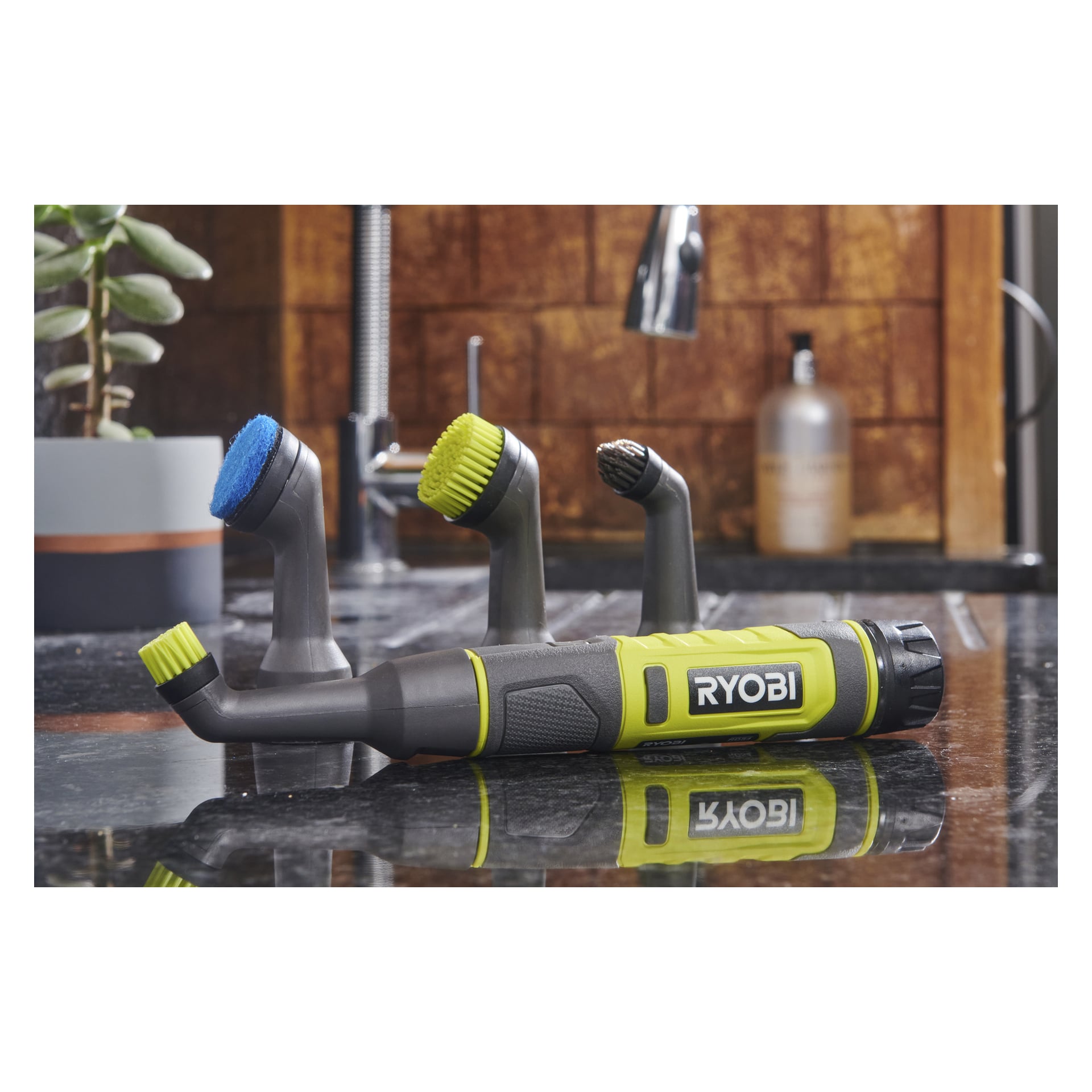 En Ryobi USB Lithium detaljrengöringsborste RDS4-0 med tre borsthuvuden står på en köksbänk nära en diskho och tvålbehållare.